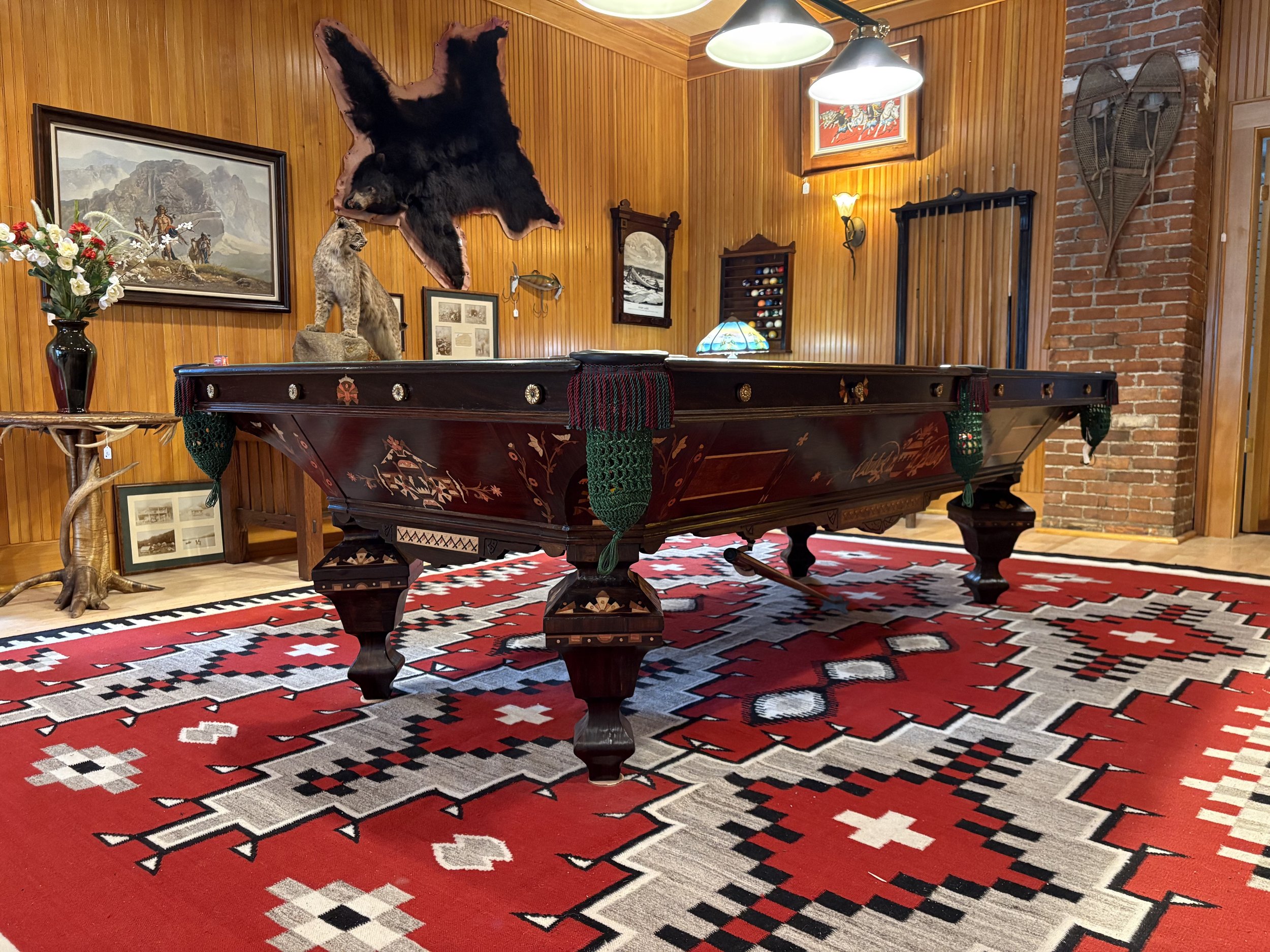 A game room with wooden-paneled walls, featuring a mounted bear hide, a mount of a mountain lion, and a pair of snowshoes on the wall. There is a pool table with green netted pockets, a striped rug with red, black, and white patterns, a floral lamp, and framed pictures and artwork on the walls.
