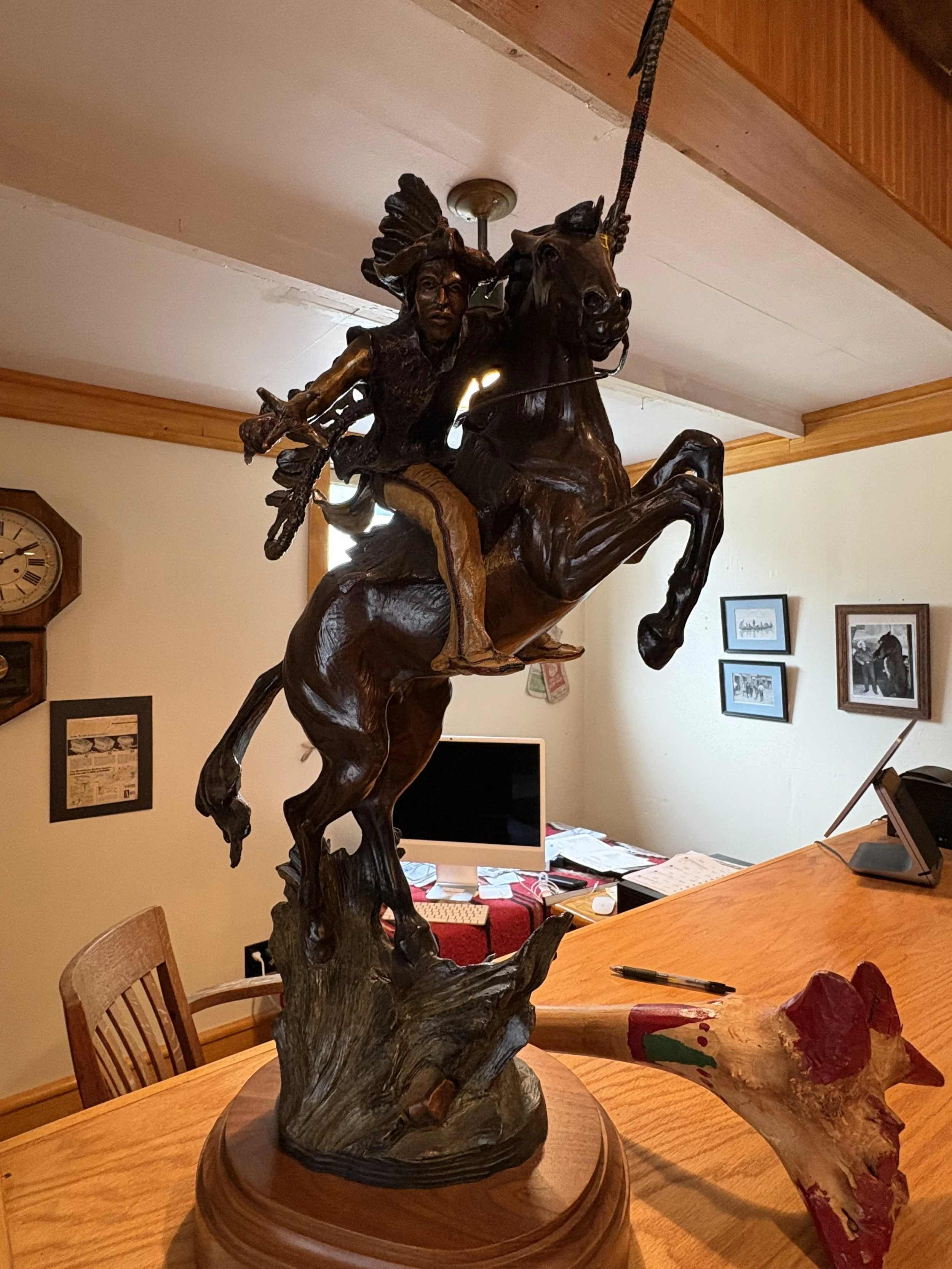 A wooden sculpture of a Native American figure riding a horse, mounted on a circular wooden base, situated on a wooden dining table within a home setting.