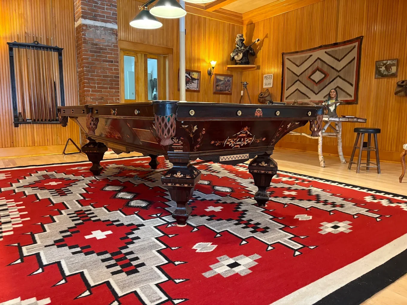 A room with wooden walls and a large red and white patterned rug, featuring an antique billiards table with decorative inlays, illuminated by ceiling lights. The room has southwestern-style artwork and a cowboy statue playing guitar on a shelf.
