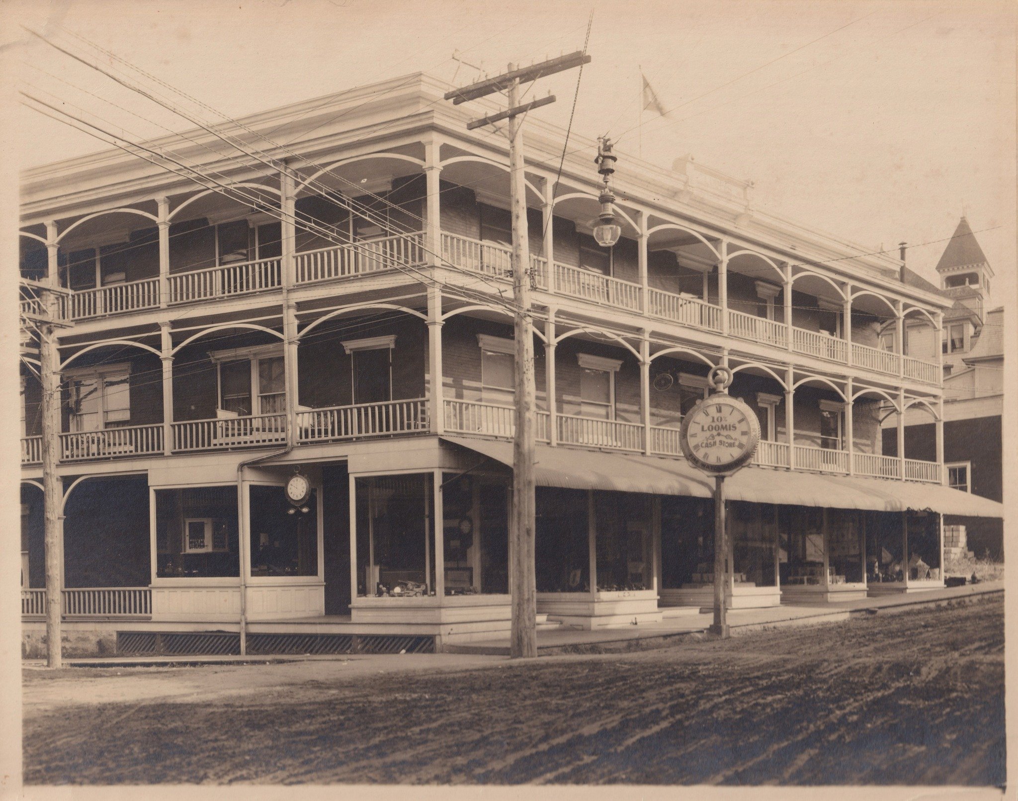 Black and white photograph of a three-story Victorian building with wraparound porches on each floor, decorative arches, and a turret with a conical roof. A utility pole and wires are in front of the building. A sign reading "Loomis Cash Store" hangs on the street level.