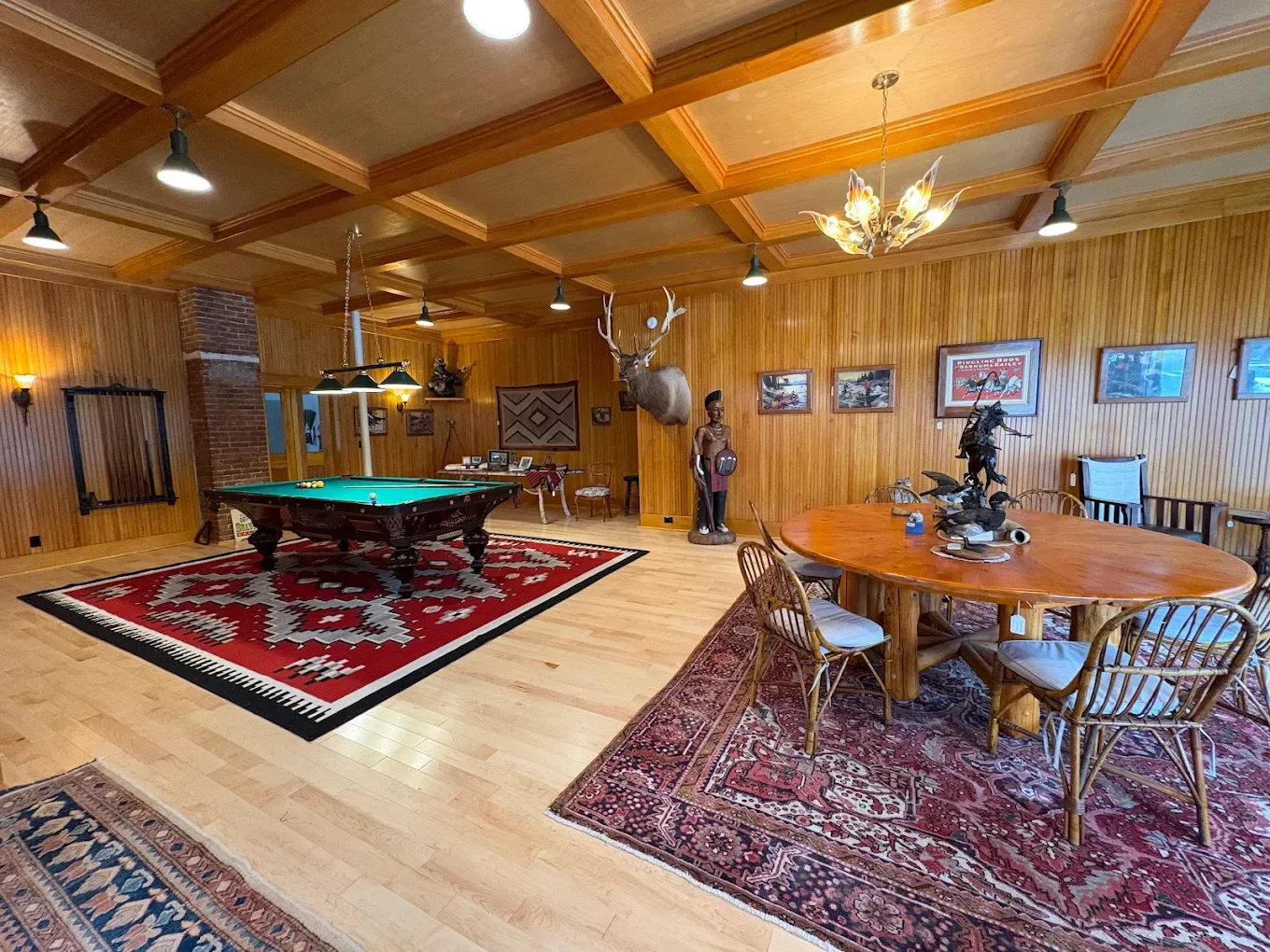 A spacious game room with wooden panel walls and ceiling, featuring a pool table on a red and white rug, a large round wooden table with chairs, a mounted deer head, decorative statues, framed artwork, and vintage-style lighting fixtures.