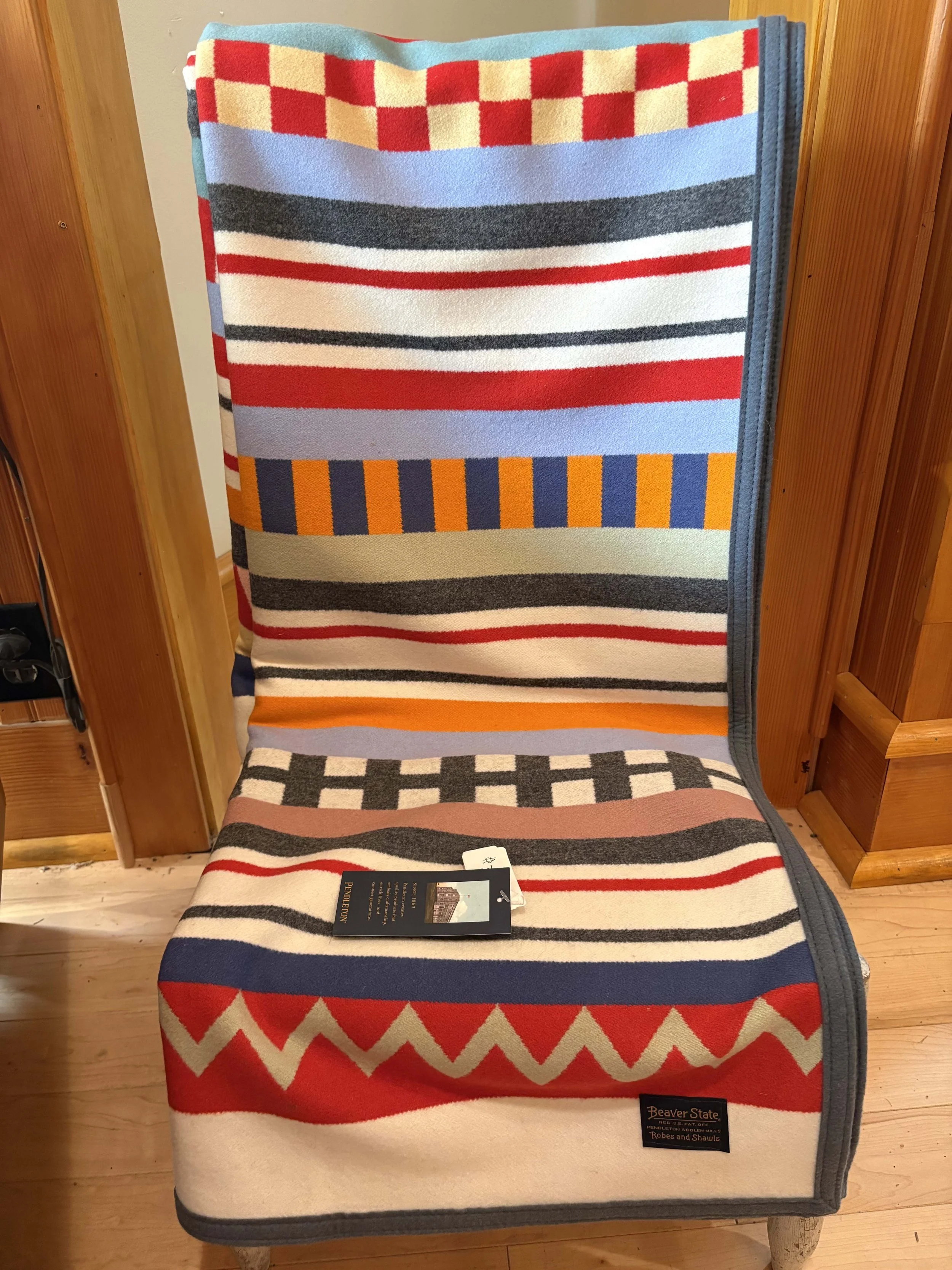 A colorful striped blanket or towel hanging over a chair with a wooden background, a black tag on the blanket reads 'Beaver State' and 'Robes and Shawls'.