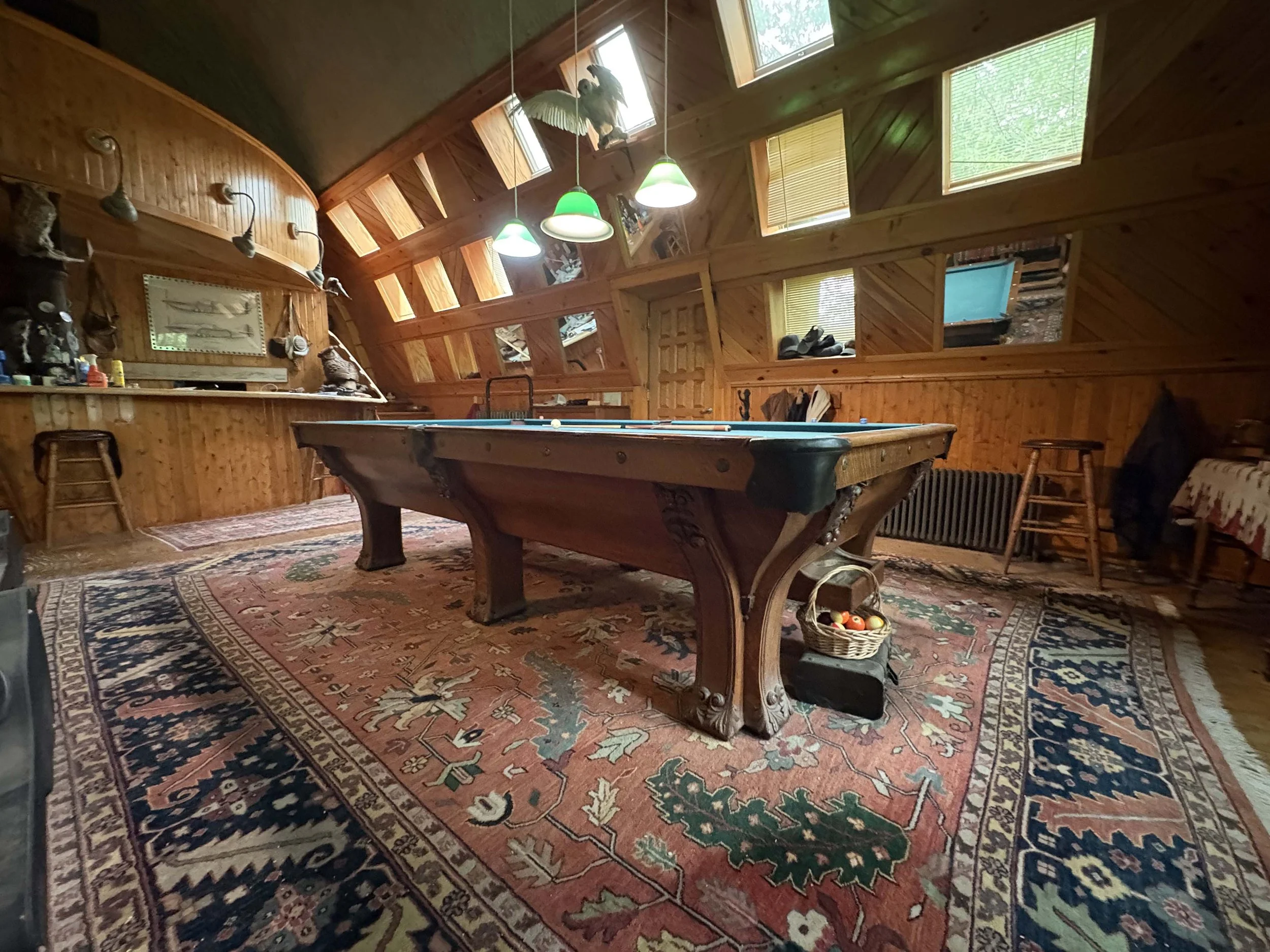 A cozy game room with wooden walls and ceiling, featuring a pool table at the center, surrounded by a patterned rug, and decorated with mounted animal skulls, fishing trophies, and a framed fish illustration. There are large windows letting in natural light, above which hang three green and white pendant lights. A basket of pool balls and other objects are also visible.
