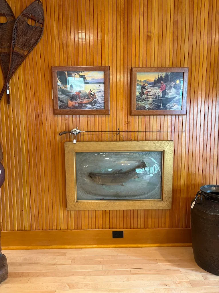 A wooden wall decorated with vintage snowshoes, two landscape paintings of people fishing in boats, and a glass case containing a large fish. There is a large ceramic pot on the floor to the right.
