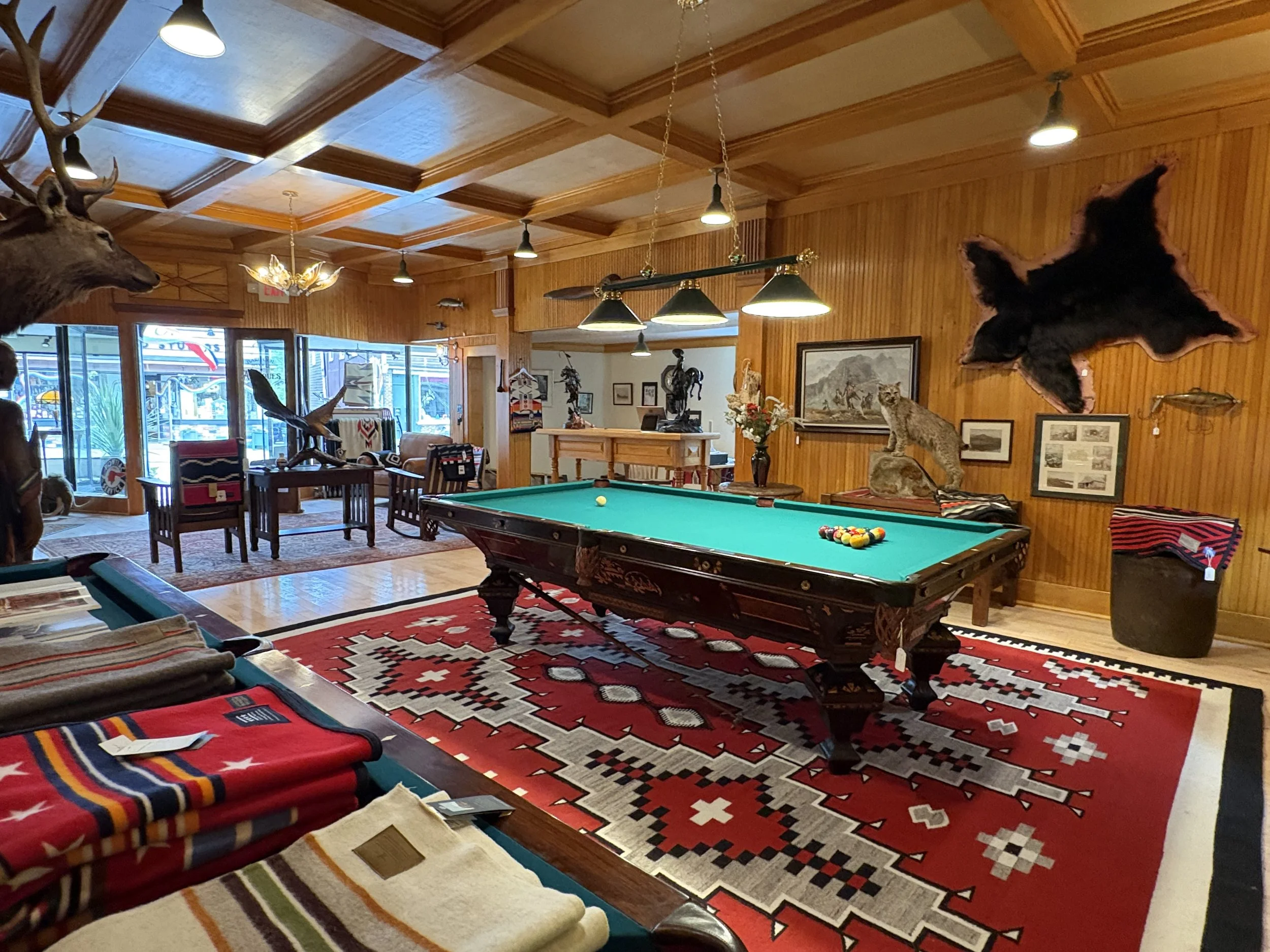 A room with wooden walls and ceiling, decorated with mounted animal heads, taxidermy animals, and framed pictures. There is a pool table in the center with pool balls and cues. The room has warm lighting and ethnic-style rugs, with several chairs and tables around.