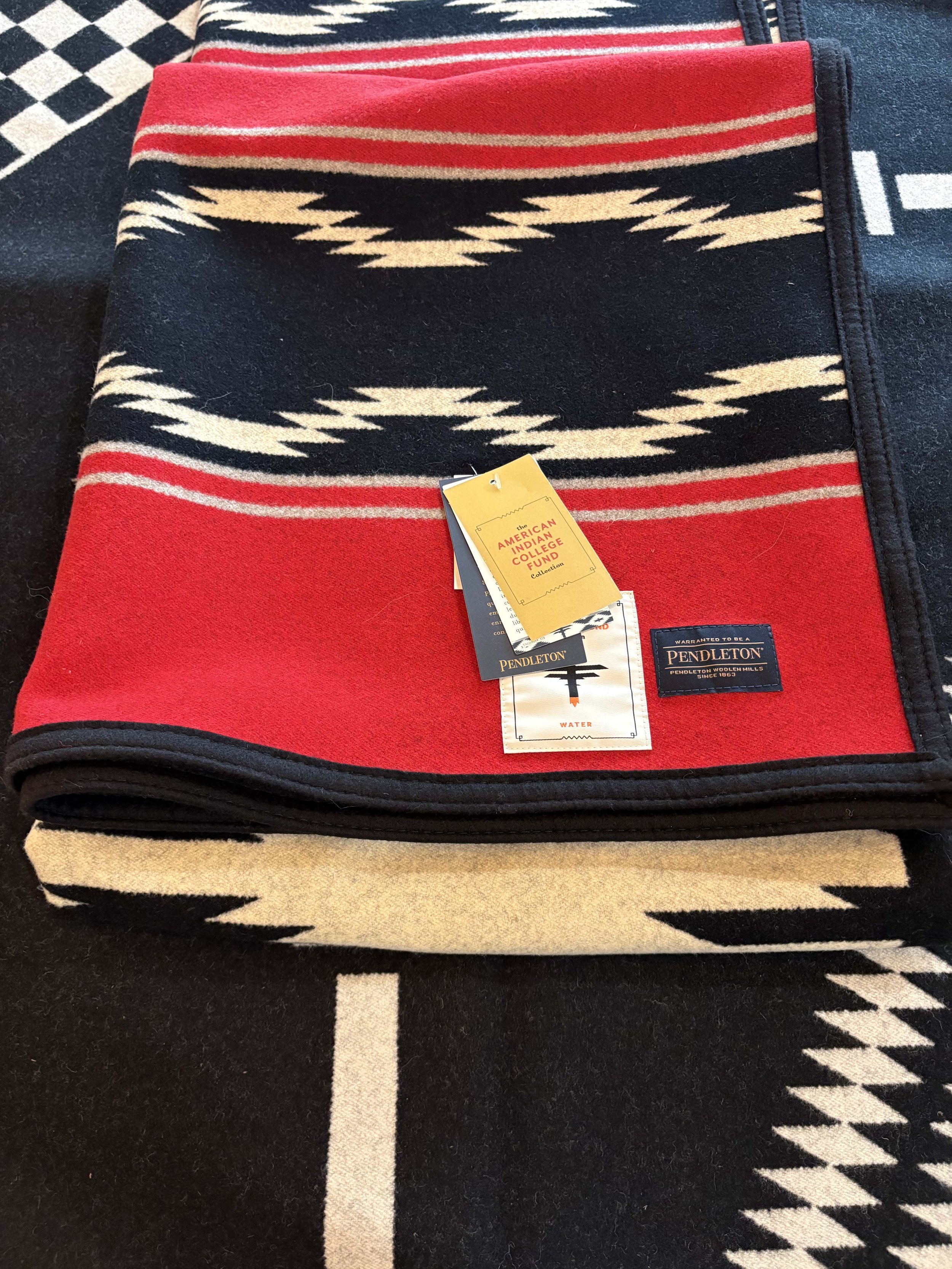A folded red and black blanket with a Navajo pattern featuring beige and black geometric shapes, placed on a black and white patterned rug.