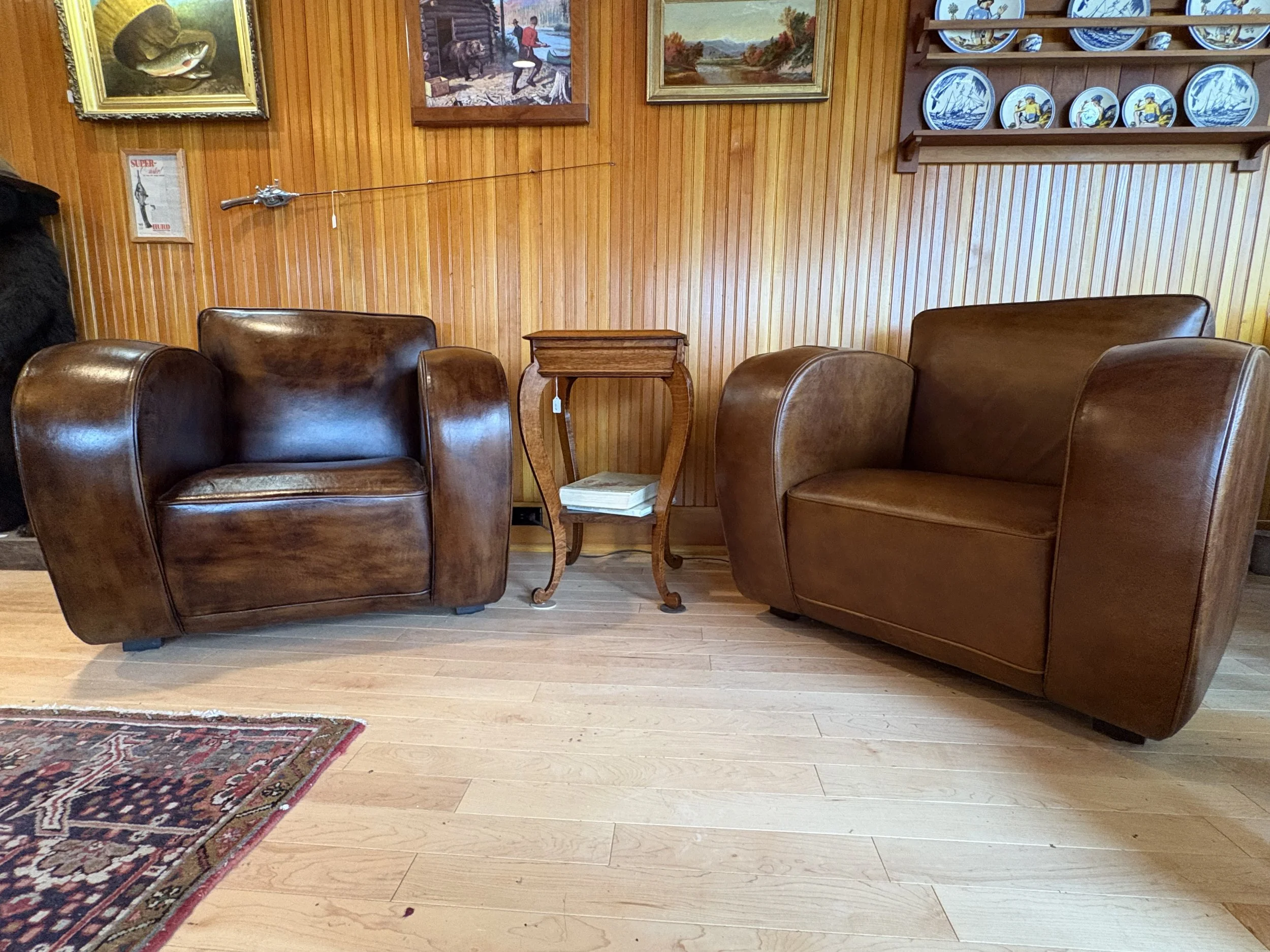 A cozy living room with wooden paneled walls, showing three brown leather armchairs, a small wooden side table with books, framed paintings, a decorative plate display shelf, and a patterned rug on a light hardwood floor.