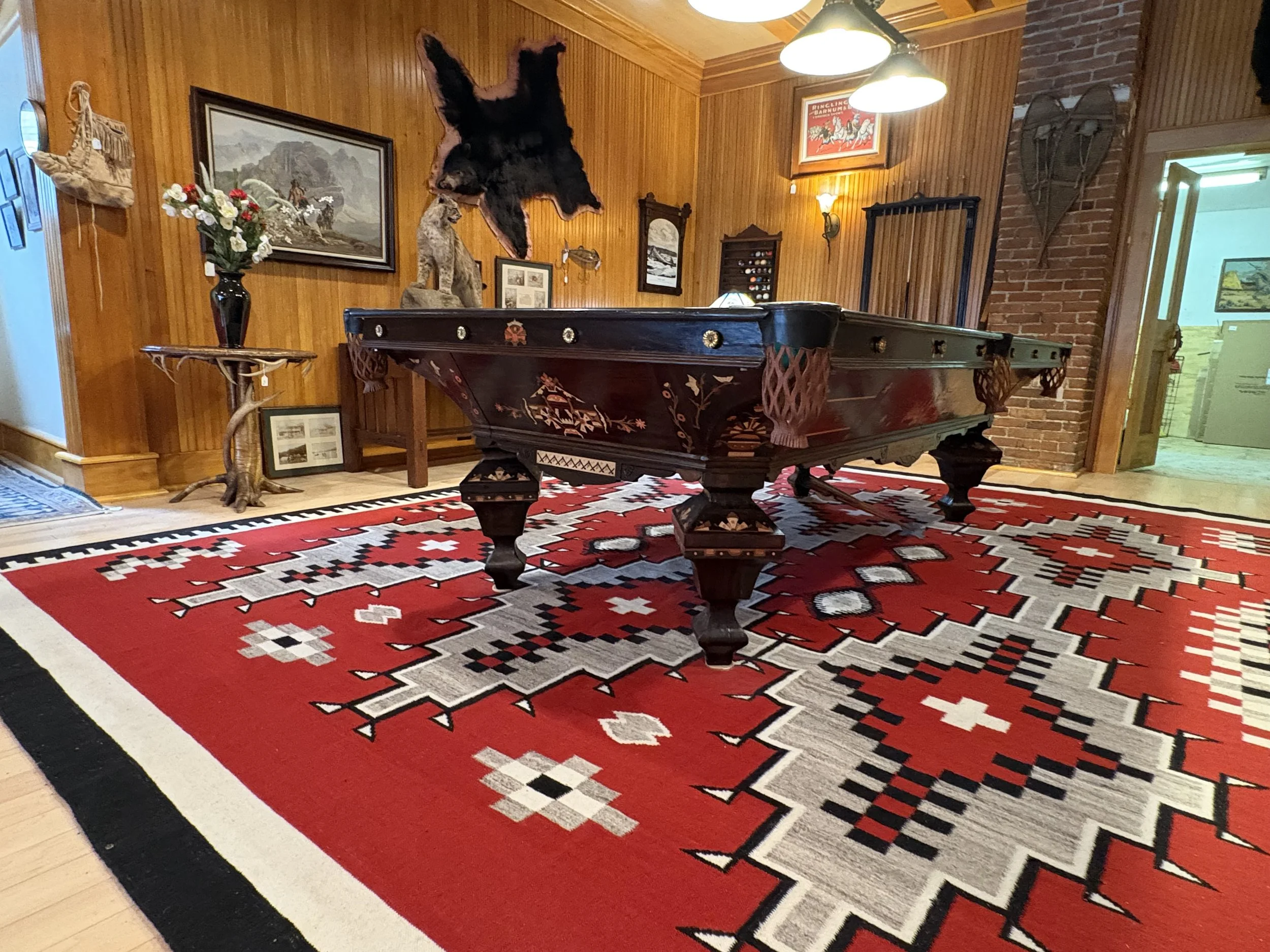 A room with wood-paneled walls and a brick wall on the right side. A decorative rug with red, black, and white patterns covers the floor. A pool table with a dark wooden finish is centered in the room. Decor items include framed pictures on the walls, a black vase with artificial flowers on a side table, a mountain landscape painting, a mounted animal pelt, and a wooden shelf with collectibles. There is a door on the right side that leads to another room, and a pair of vintage skis hanging on the brick wall.