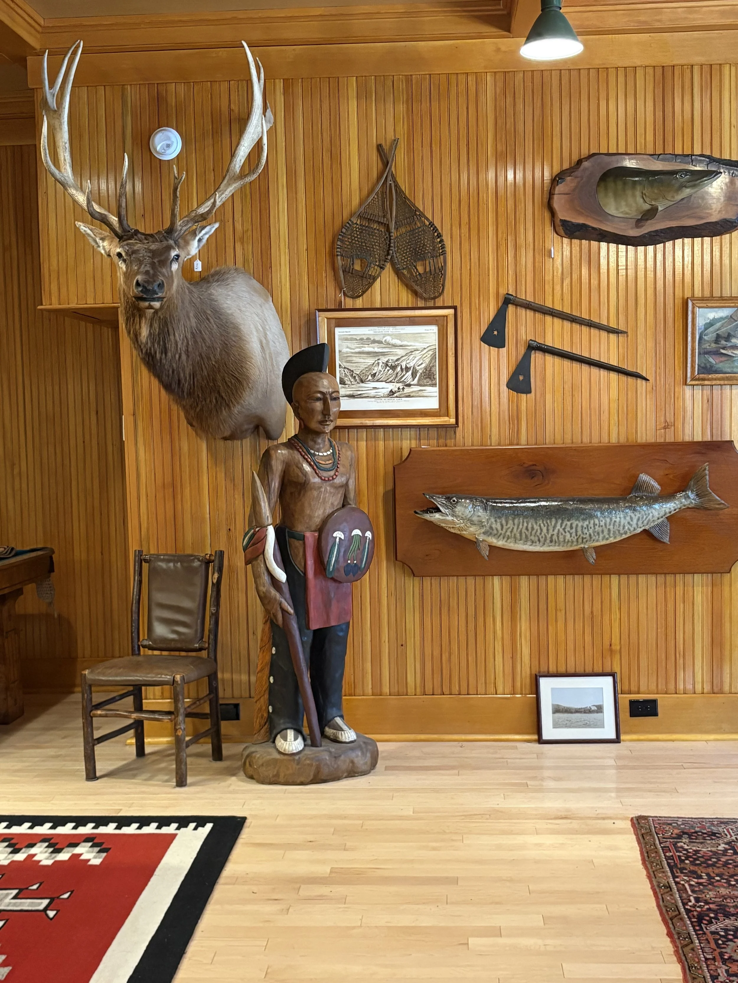 A rustic game room with wood-paneled walls and ceiling, featuring a pool table on a patterned red and white rug, a round dining table with wicker chairs, a mounted deer head on the wall, and various decorative items including a wooden sculpture and framed pictures.