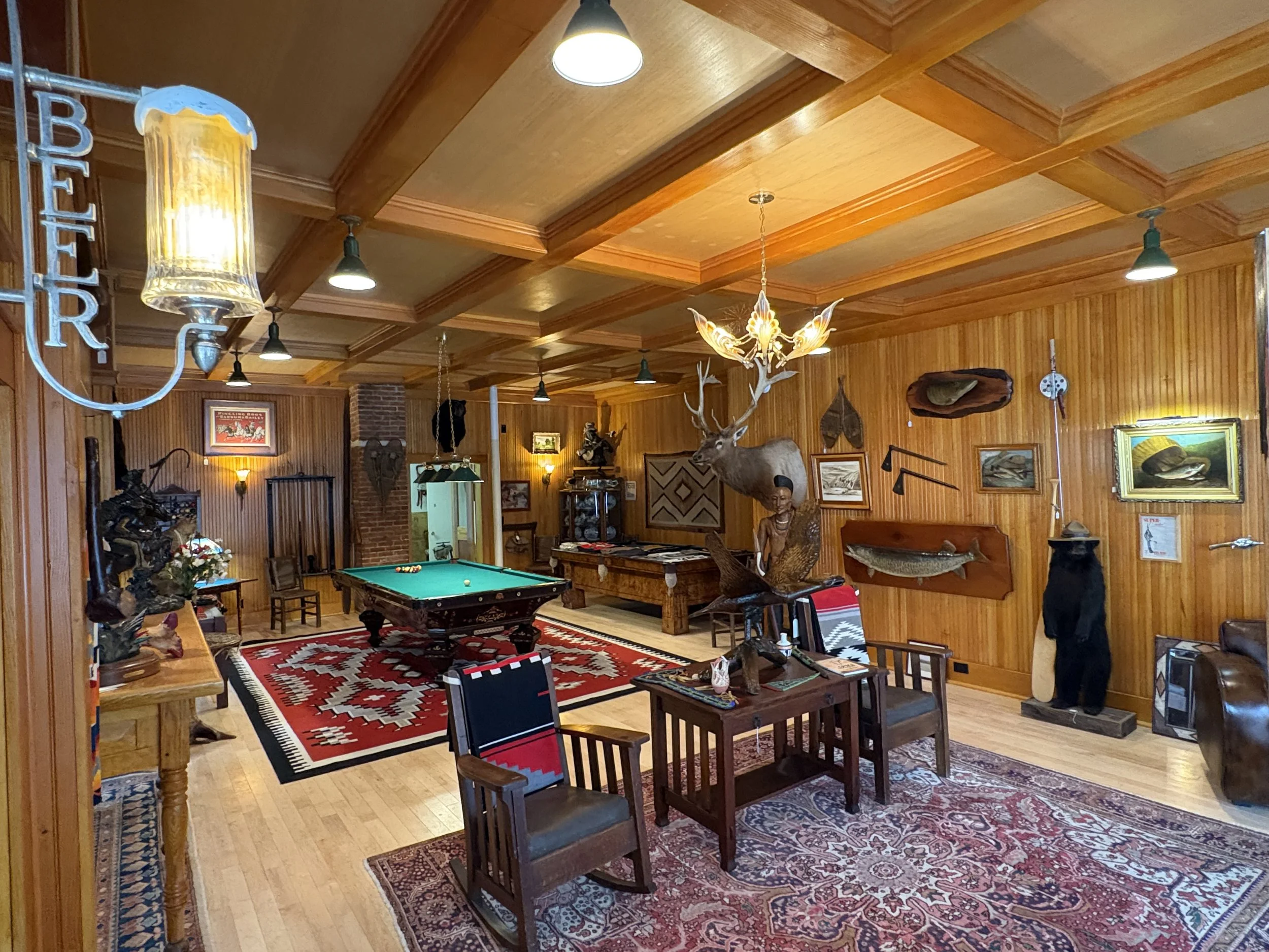The image shows a cozy, rustic interior with wood-paneled walls and ceiling, decorated with various artwork, framed pictures, and taxidermy, including a large mounted deer head. There is a pool table in the center, surrounded by chairs and tables with books and decorative items on top. Several pendant lights hang from the ceiling, and a colorful southwestern-style rug covers part of the wooden floor.