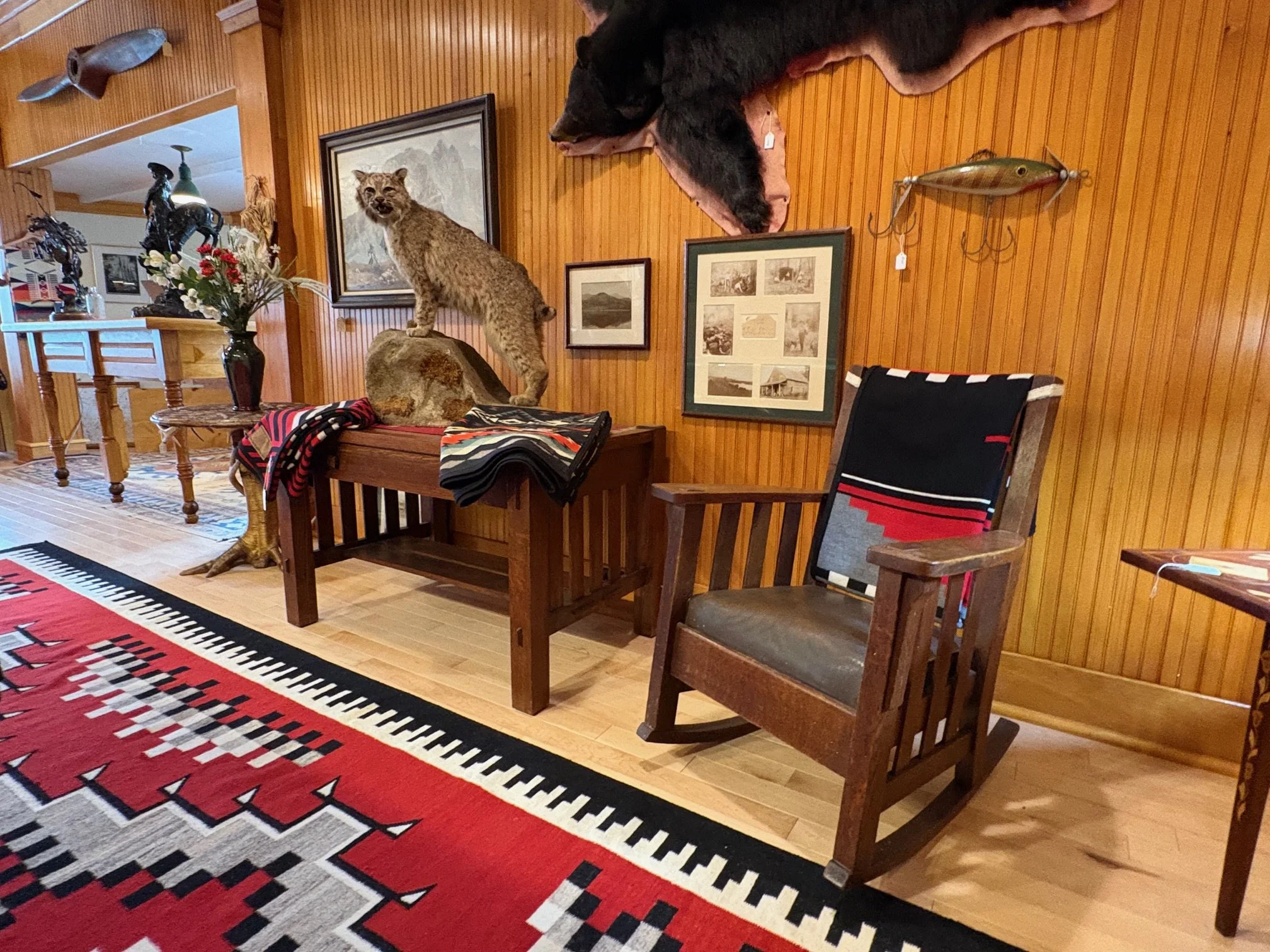 A room with wood-paneled walls decorated with animal-themed artwork and mounted animal hides. A taxidermy cougar is standing on a rock in the center of the room. The room has a wooden table with a vase of flowers, a black lamp, and a striped throw blanket. Wooden chairs and a patterned area rug are also visible.