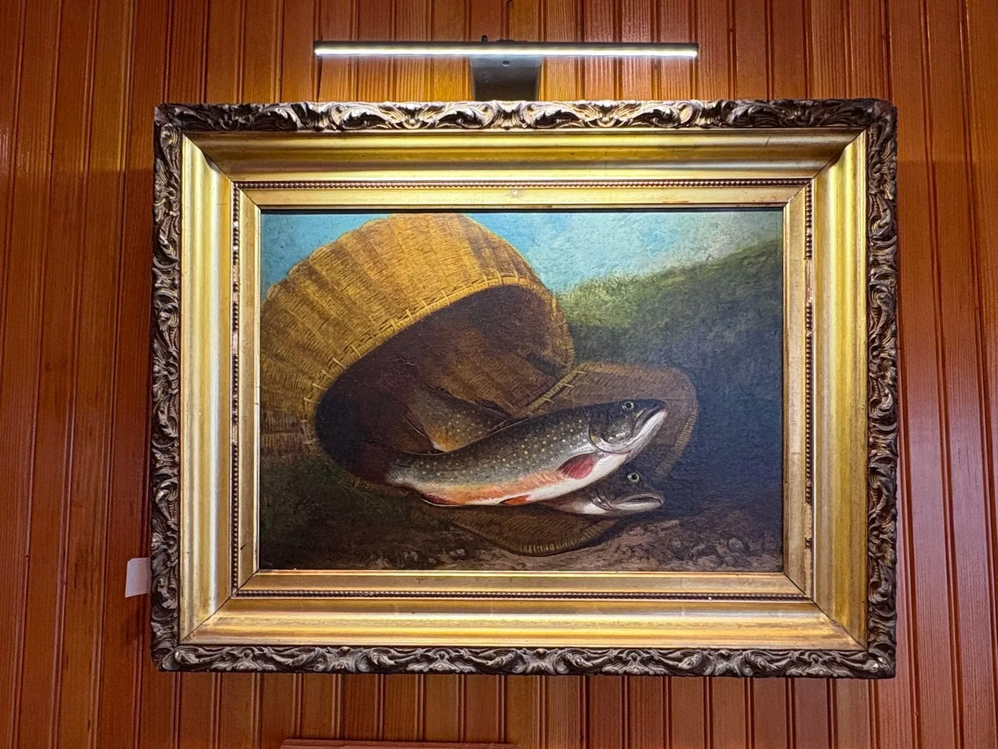 A painting of a fish with a mountain in the background, framed with an ornate border, hung on a wooden wall.