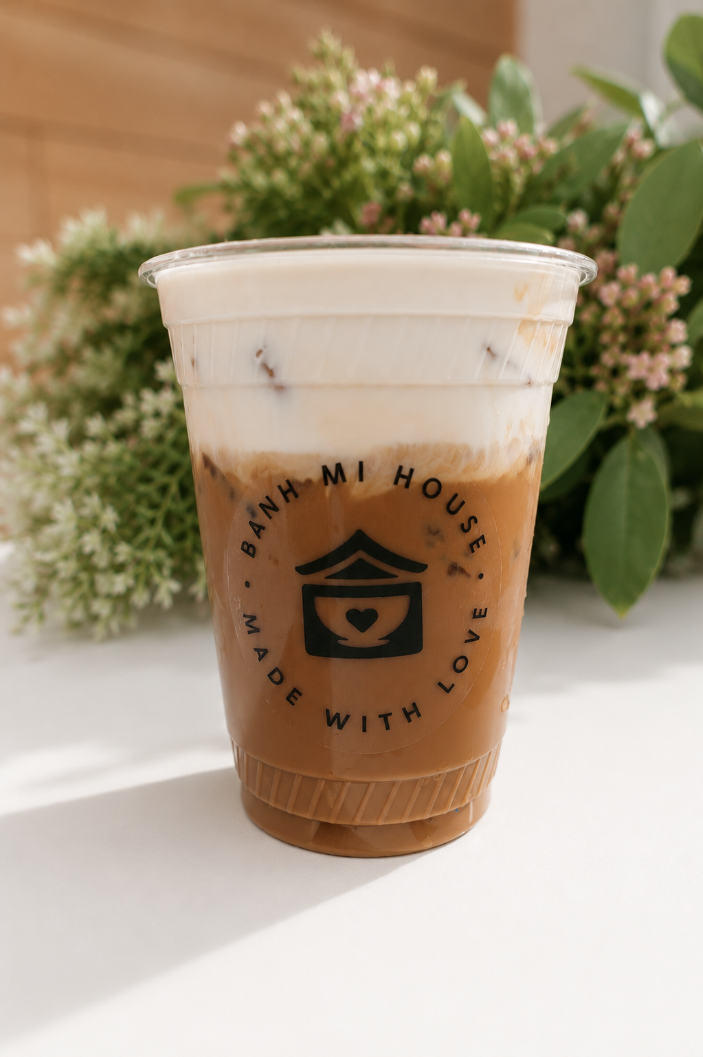 A plastic cup of iced coffee with milk, featuring a black logo and text saying 'Banh Mi House Made with Love,' placed on a white surface with green foliage in the background.