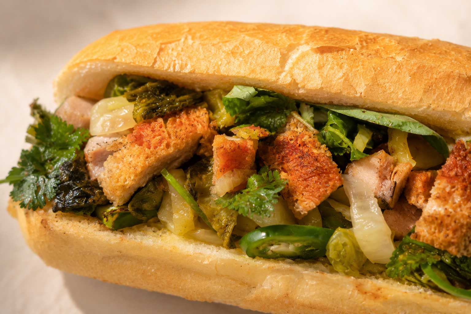 Close-up of a sandwich filled with fried chicken, leafy greens, herbs, and sliced chili peppers on a baguette-style bread.