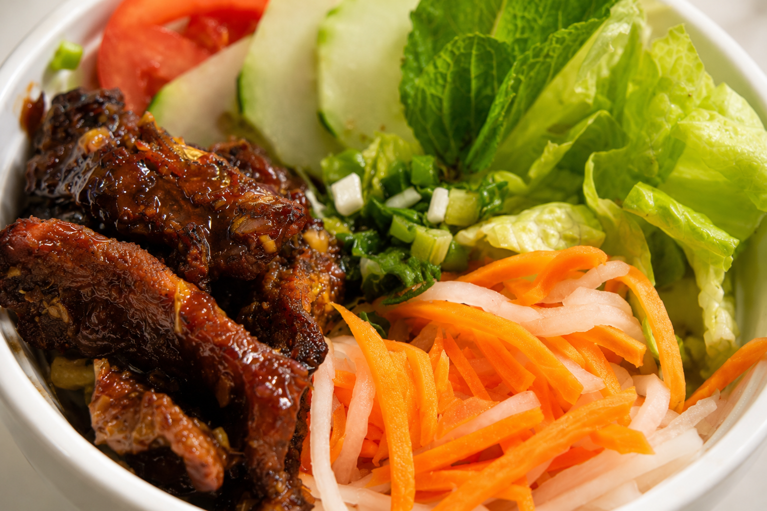 A close-up of a bowl containing grilled barbecue chicken, fresh vegetables including sliced tomato, cucumber, lettuce, chopped green onions, and shredded carrots and daikon radish.