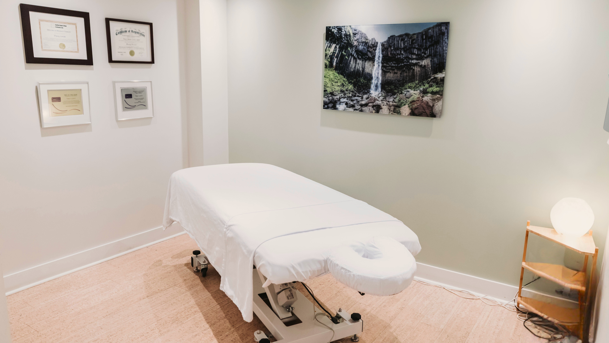 A massage table covered with a white sheet in a room with framed certificates on the wall and a large wall art picture of a waterfall.