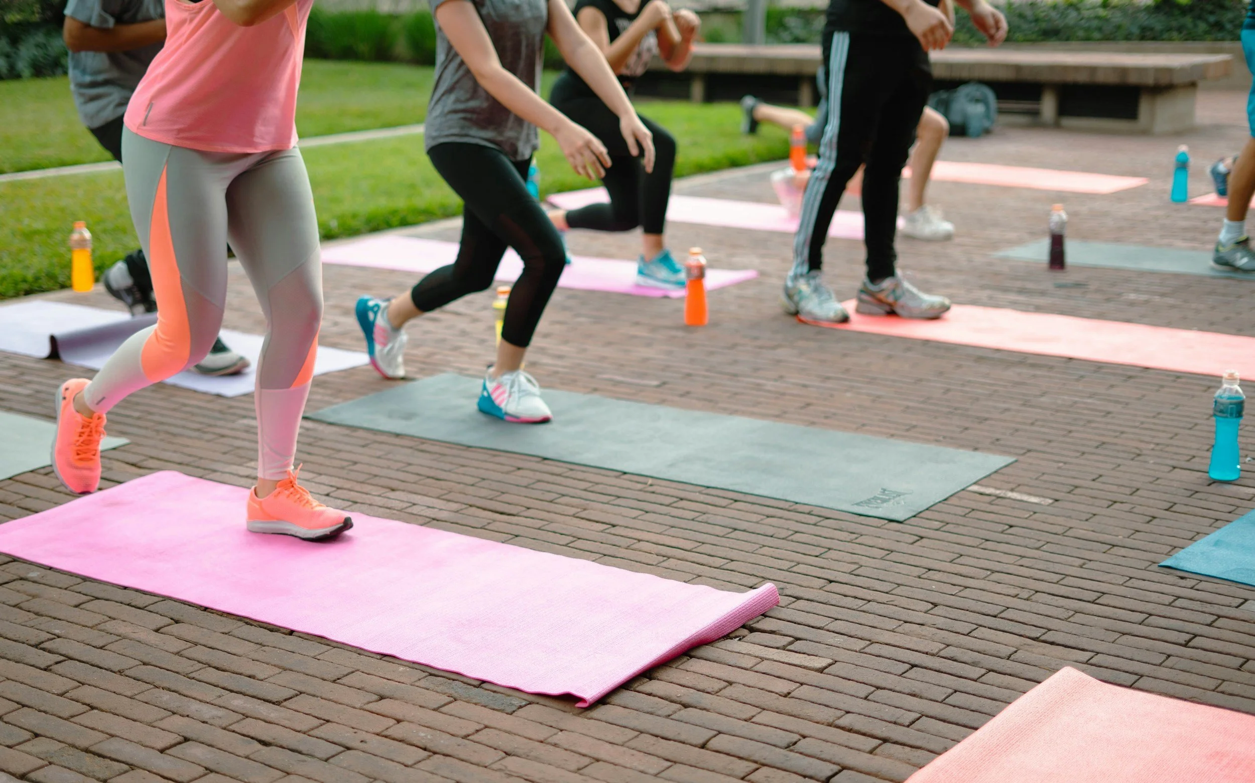 Group of people participating in an outdoor yoga or fitness class on colorful mats, wearing athletic clothing, with water bottles placed next to them on the brick pavement.