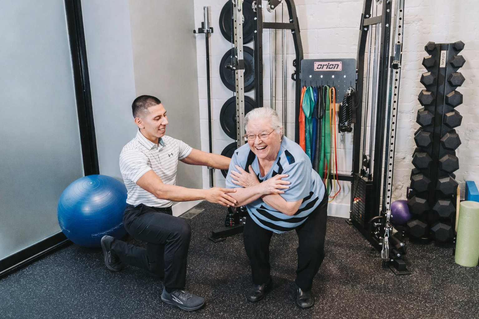 A young male fitness trainer helping an elderly woman laugh during a workout session in a gym, with workout equipment in the background.