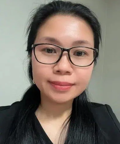 A woman with glasses and long black hair taking a selfie in a plain indoor setting.