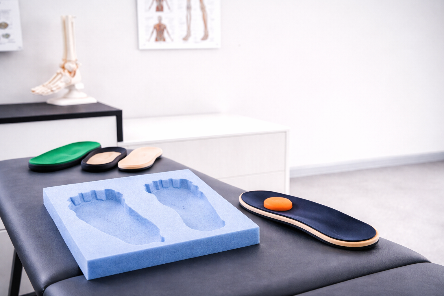 Medical podiatry tools arranged on a black examination table, including a foam foot mold, two orthotic insoles (green and beige), a black insole, and a black orthotic with an orange ball on top, with anatomical charts and models in the background.