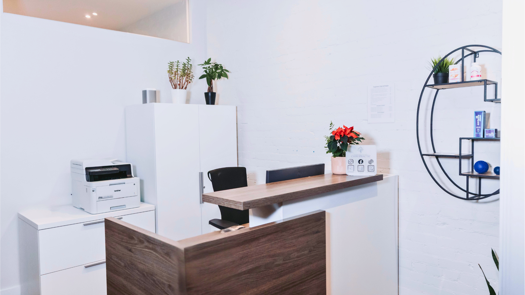 Reception area with a wooden counter, white cabinets, potted plants, a poinsettia, a chair, and a black metal shelf on the wall holding bottles and books.