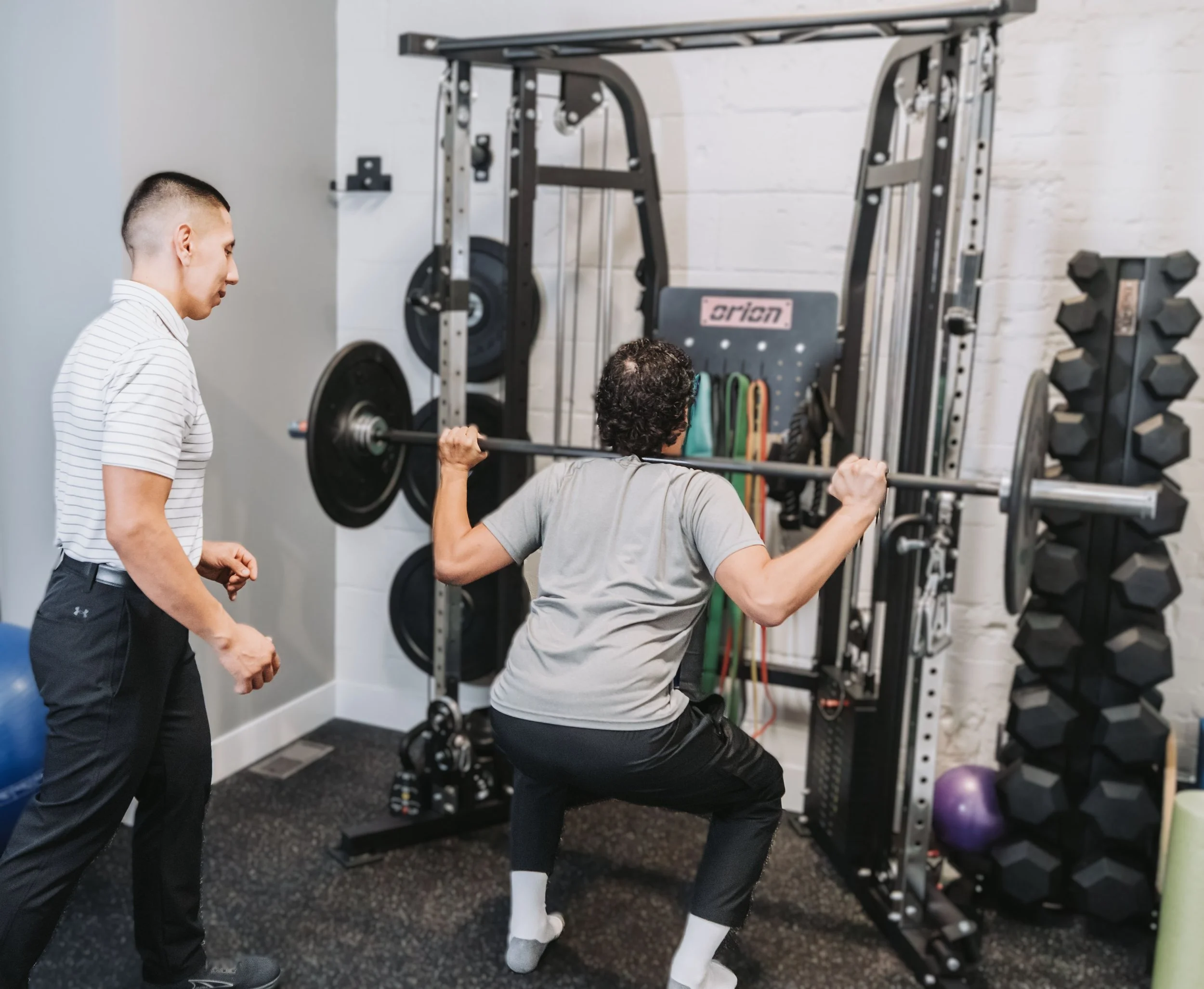 A person performing a squat exercise with a barbell on their shoulders, assisted by a spotter, in a gym setting with weight racks and exercise equipment in the background.