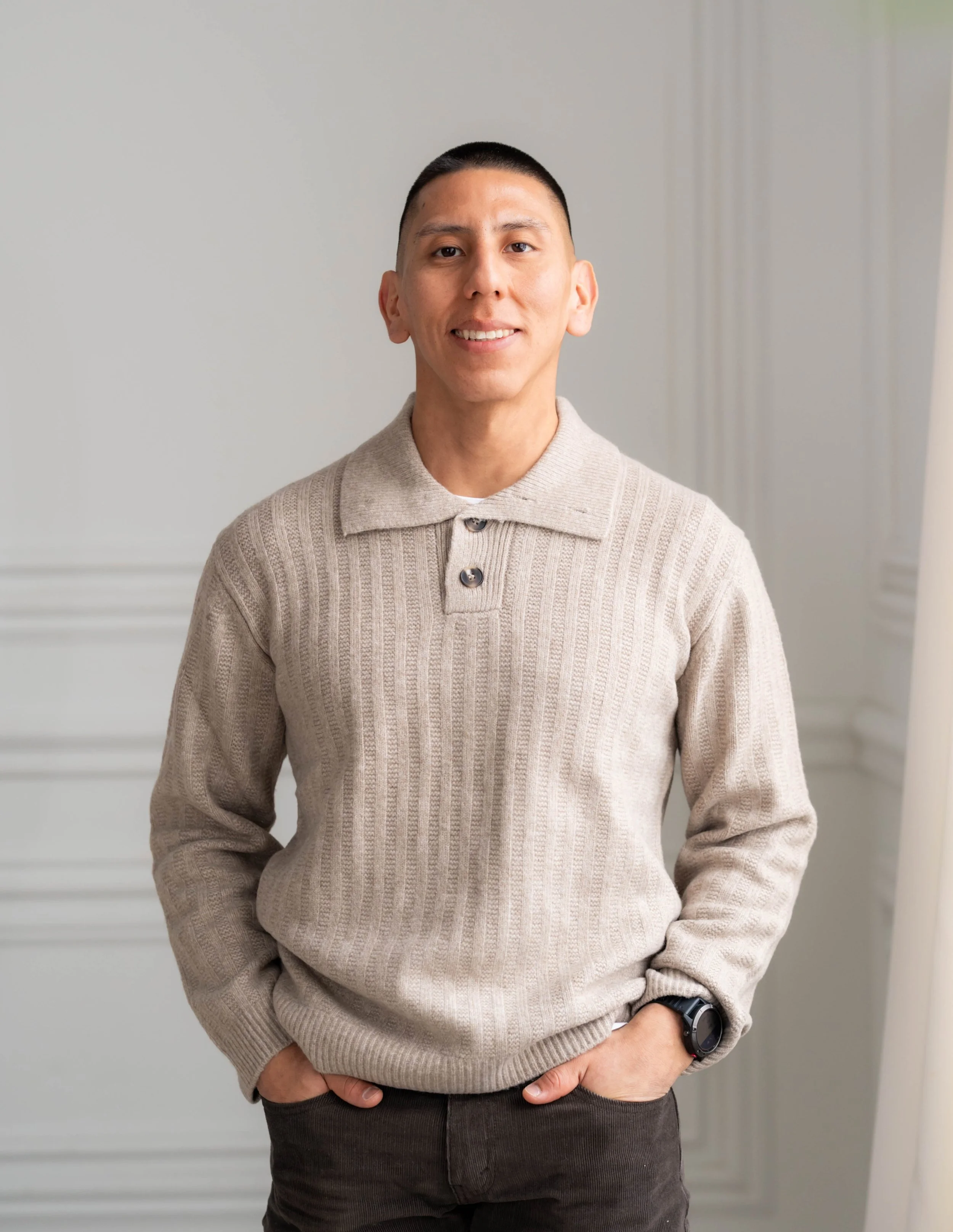 A man standing indoors wearing a beige knitted sweater over a collared shirt. He has short black hair, a slight smile, and is wearing a black wristwatch. His left hand is resting in his pocket.