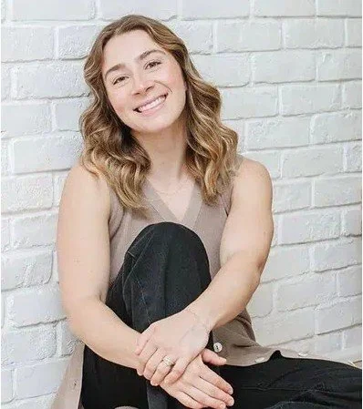 A young woman with wavy brown hair sitting against a white brick wall, smiling, wearing a beige sleeveless top and black pants.