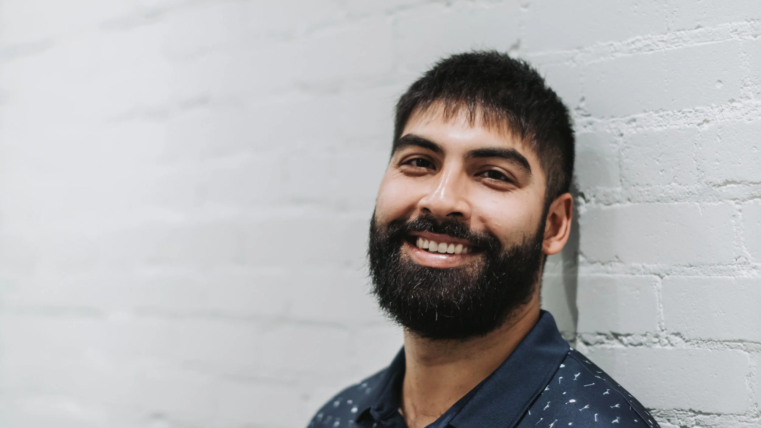 Smiling man with dark hair and beard leaning against a white brick wall, wearing a dark shirt with white patterns.