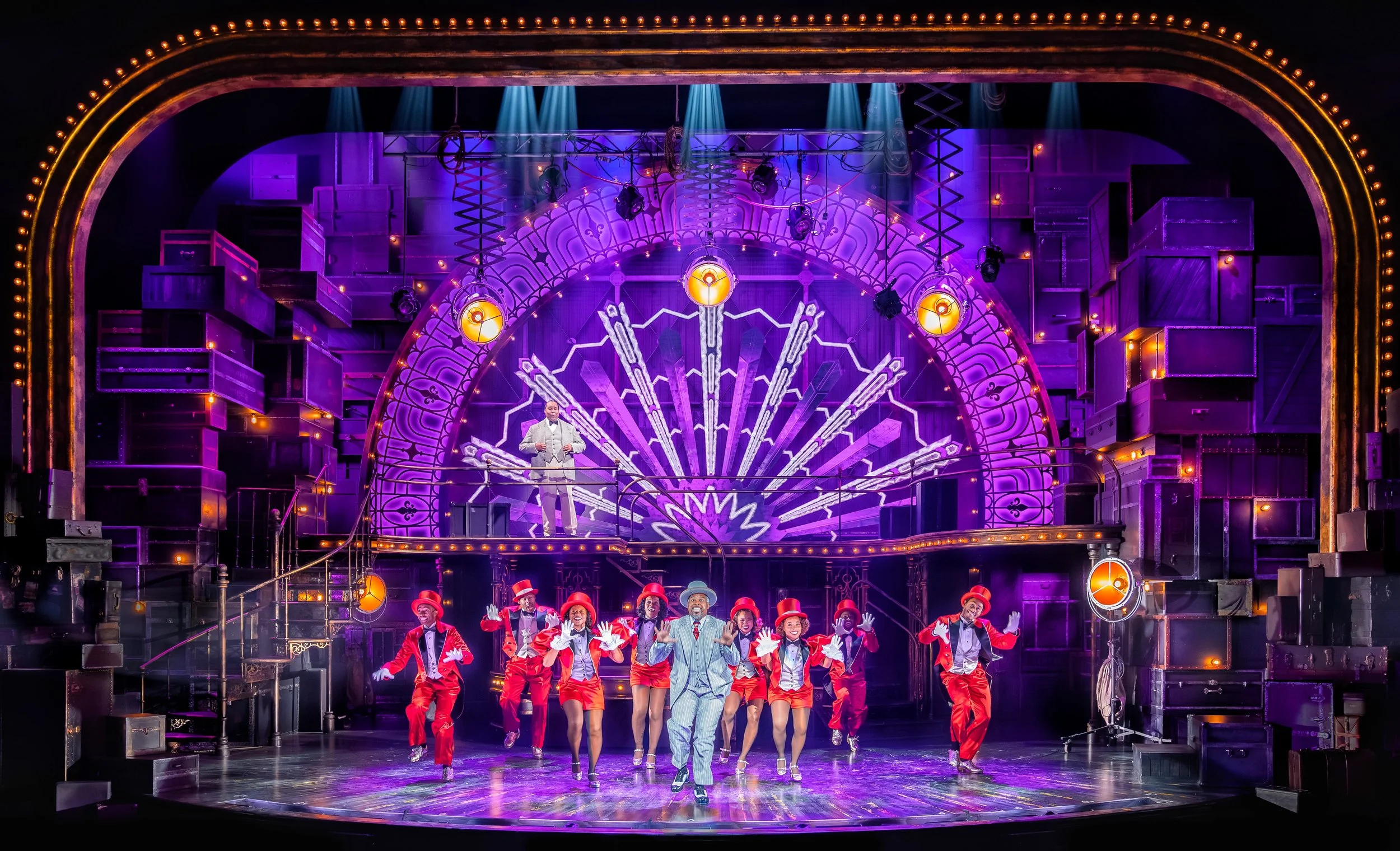 A theatrical stage performance with a group of dancers in red costumes and a man in a striped suit at center, under vibrant purple and yellow lights with an art deco backdrop.