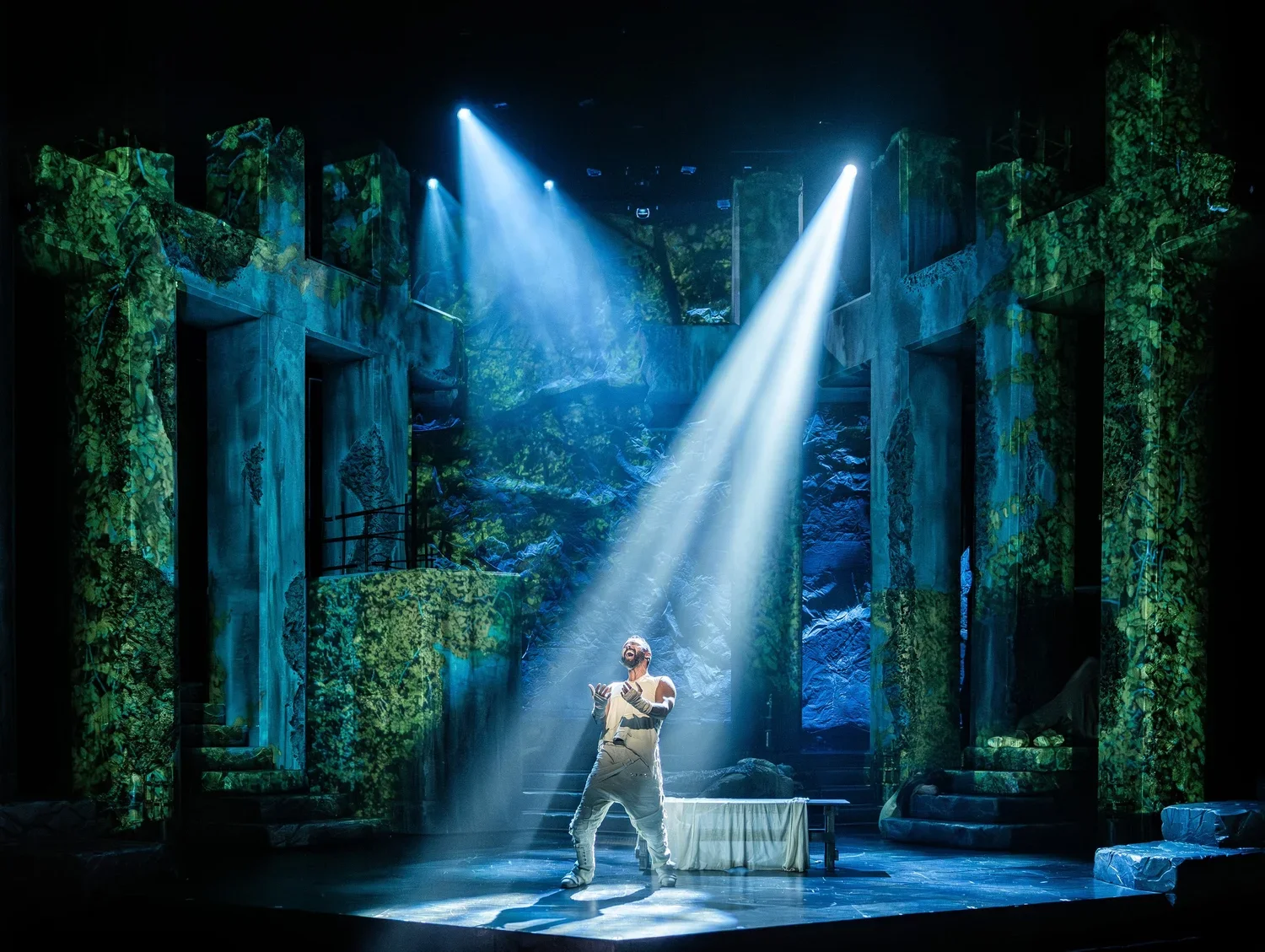 A theatrical stage with lush green foliage background, dramatic lighting, and a performer in costume singing or acting, illuminated by a spotlight.
