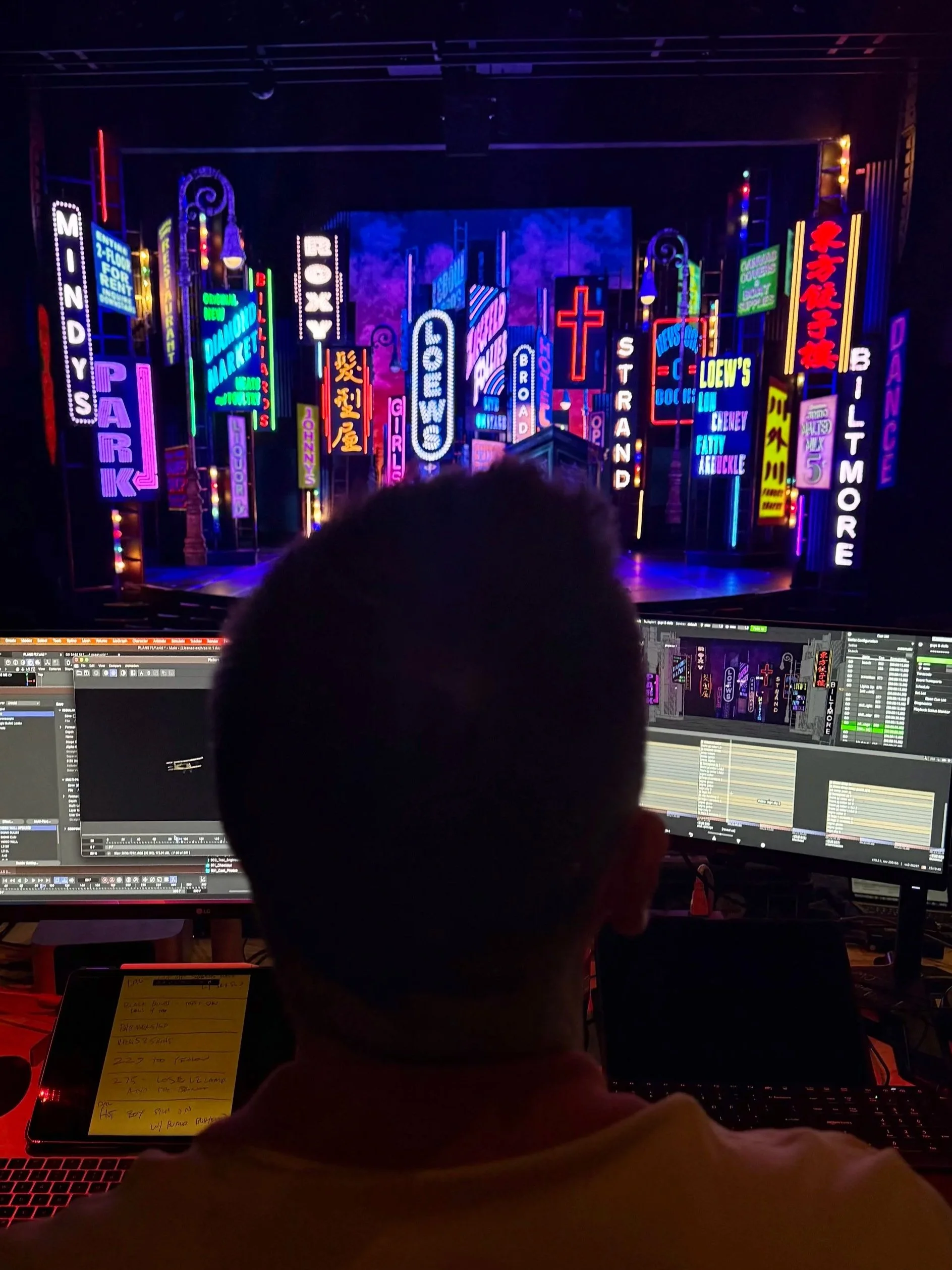 A person working on a computer in a dark room with a large screen displaying vivid neon signs of city billboards and signs.