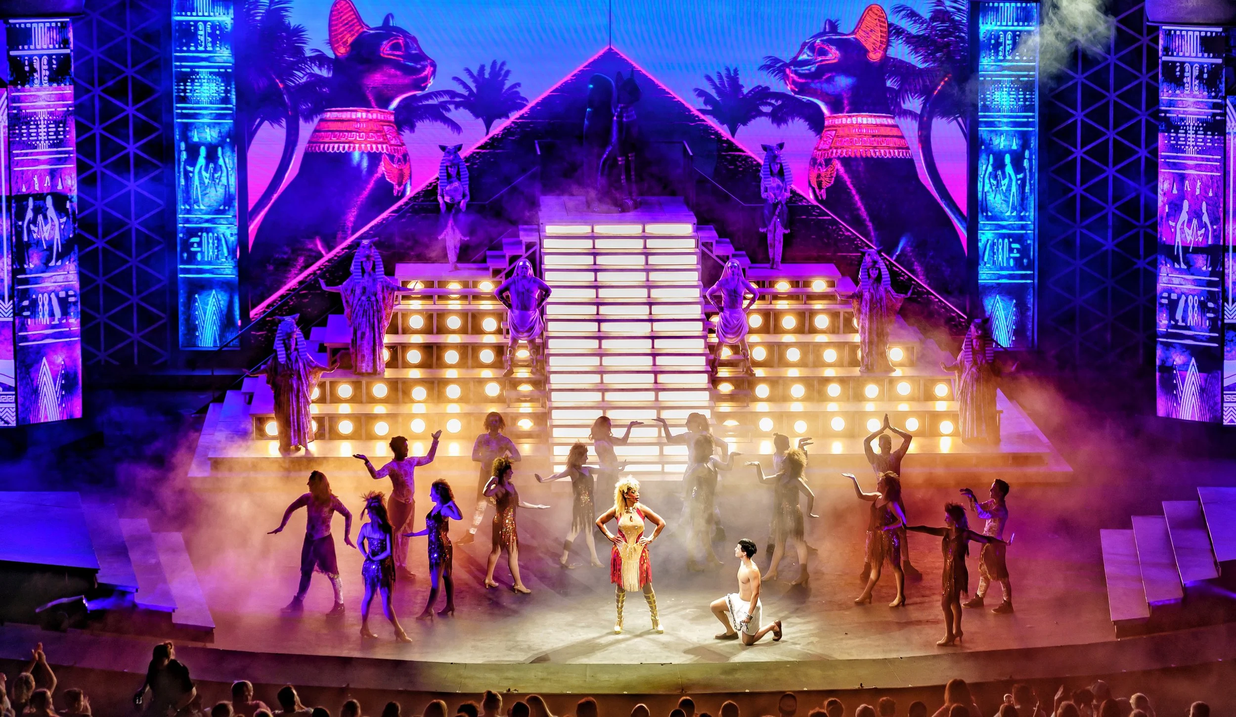 Theatrical stage performance with a large Egyptian-themed set, including pyramids and Egyptian statues. Dancers in colorful costumes perform in front of an elaborate backdrop with neon lighting and smoke effects.