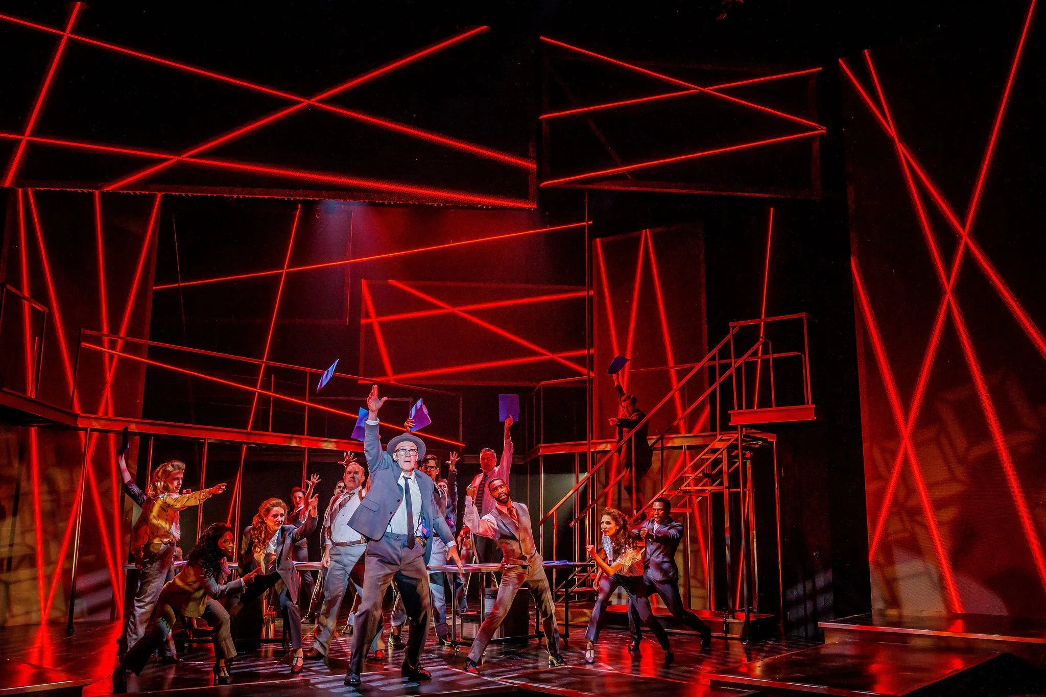 A theater stage scene with multiple actors dressed in formal and business attire, some raising their hands or holding papers, with a modern, illuminated red laser light set design in the background.