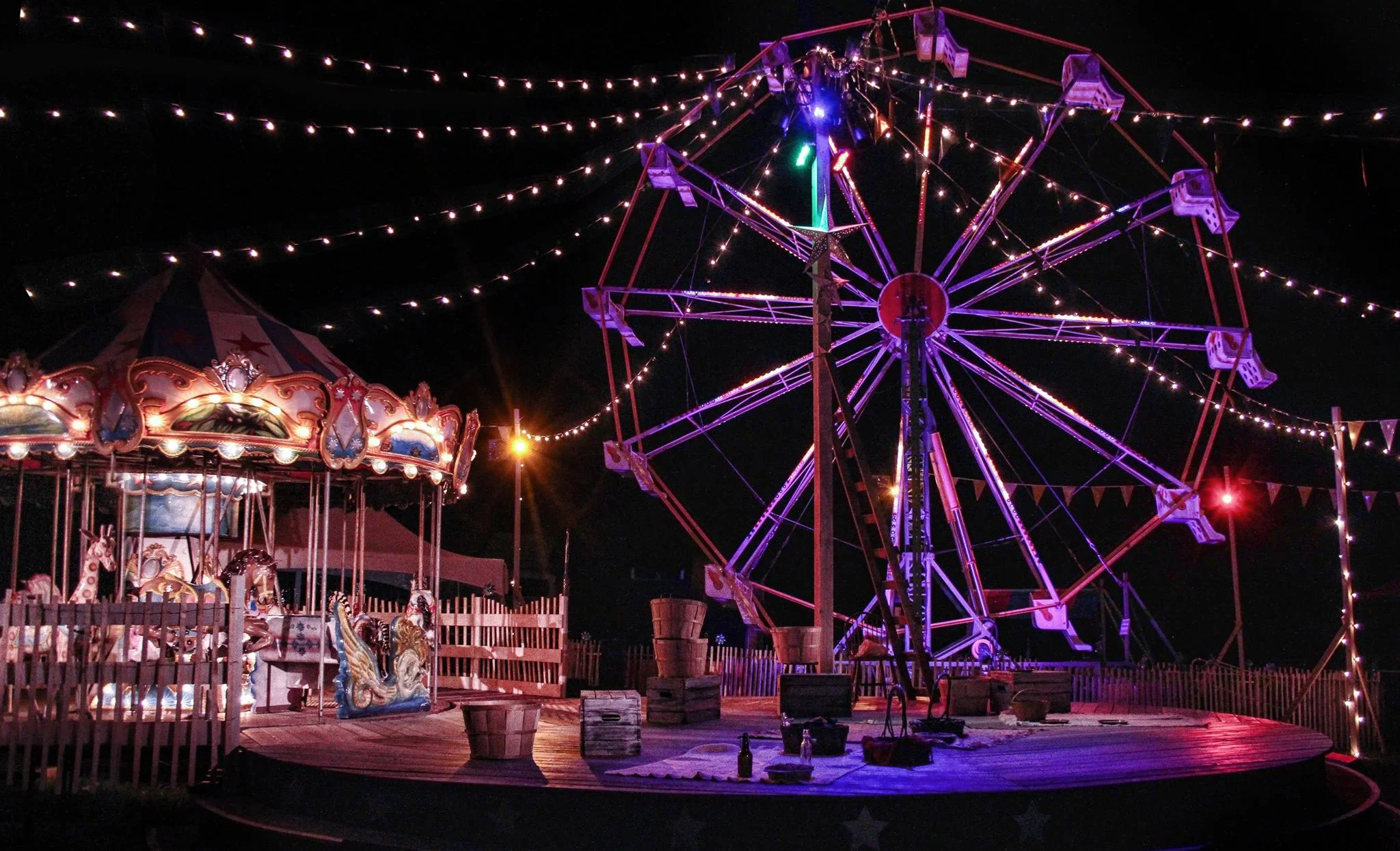 Empty amusement park rides at night, including a carousel with animal figures and a large Ferris wheel with colorful lights, surrounded by string lights and a fence.