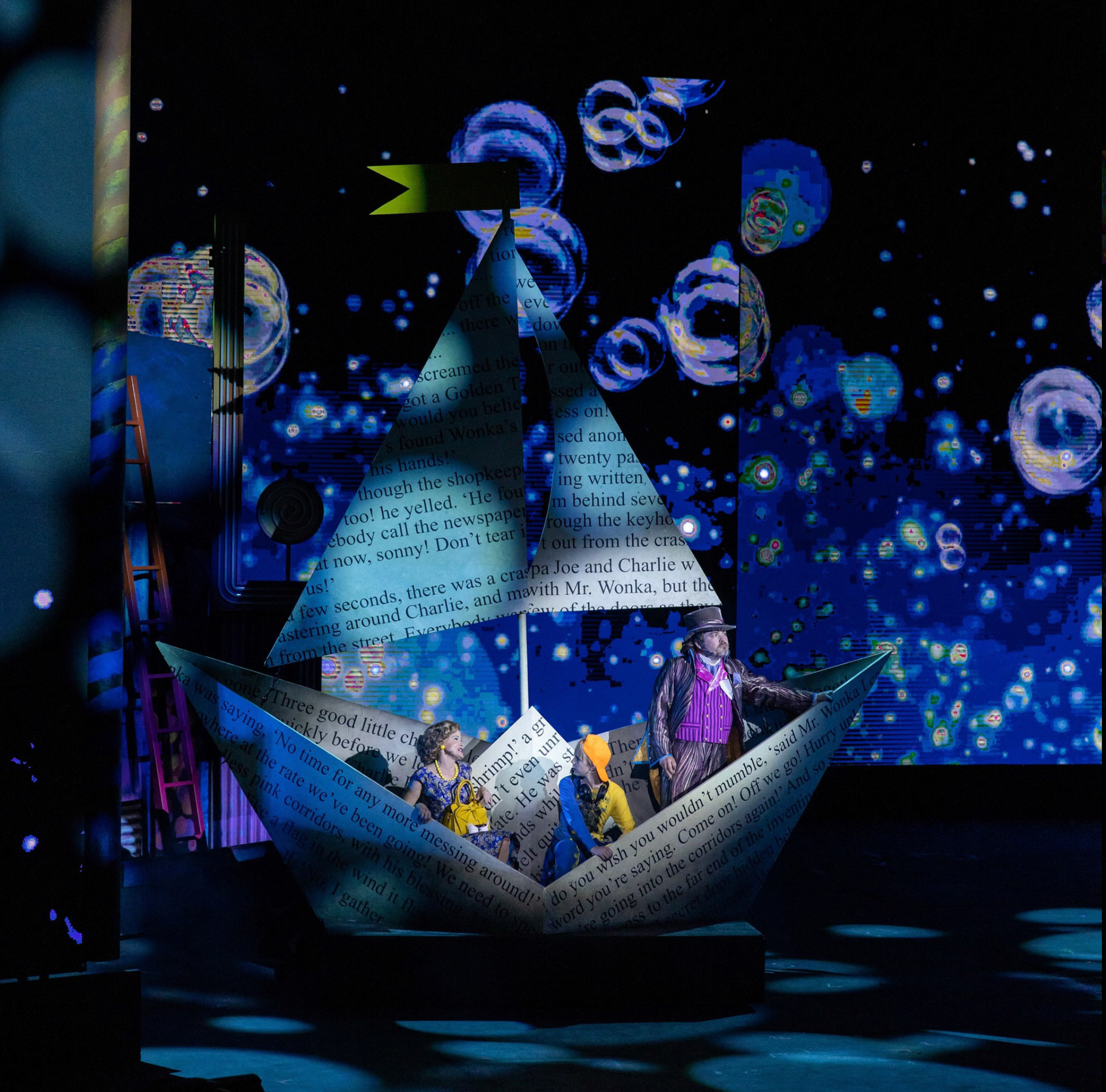 Stage scene from 'Willy Wonka & the Chocolate Factory' with characters in a paper boat set, large digital backdrop of bubbles and stars, and a boat-shaped platform.