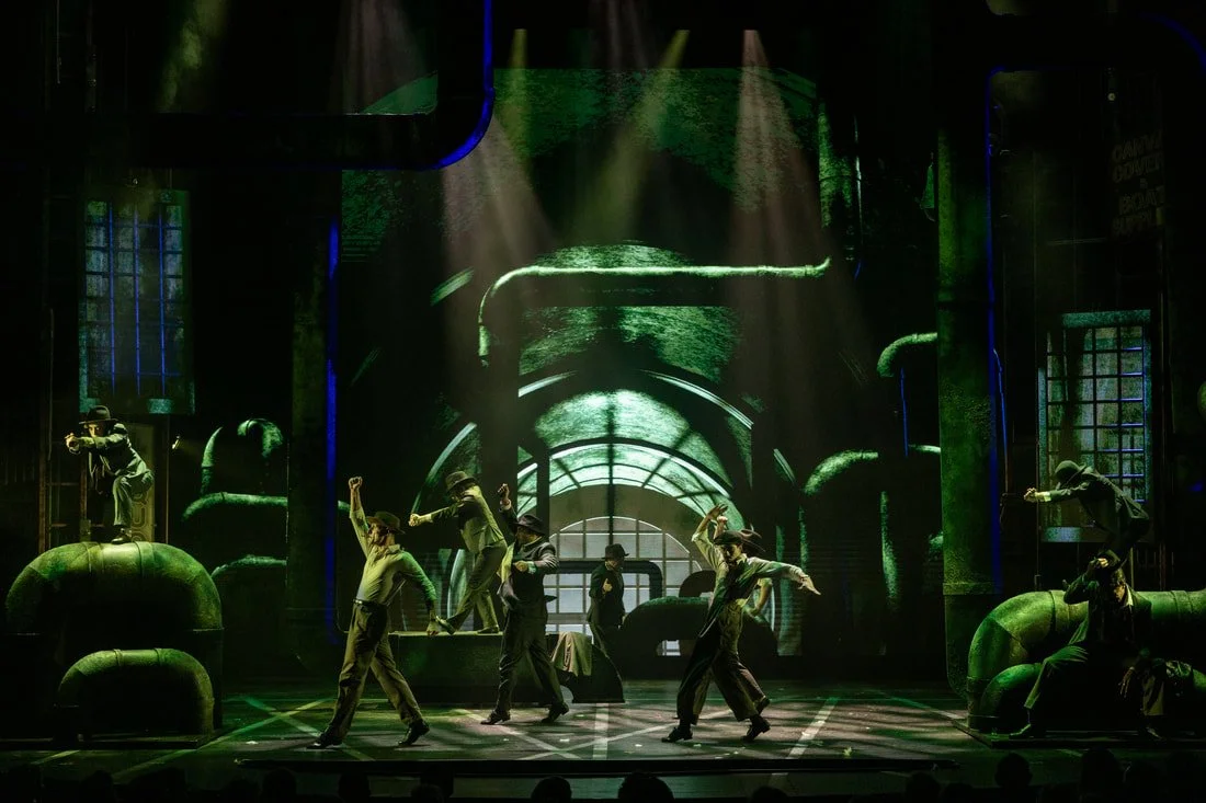 Stage performance with dancers in suits and hats, green industrial background with pipes and windows, and dramatic lighting.