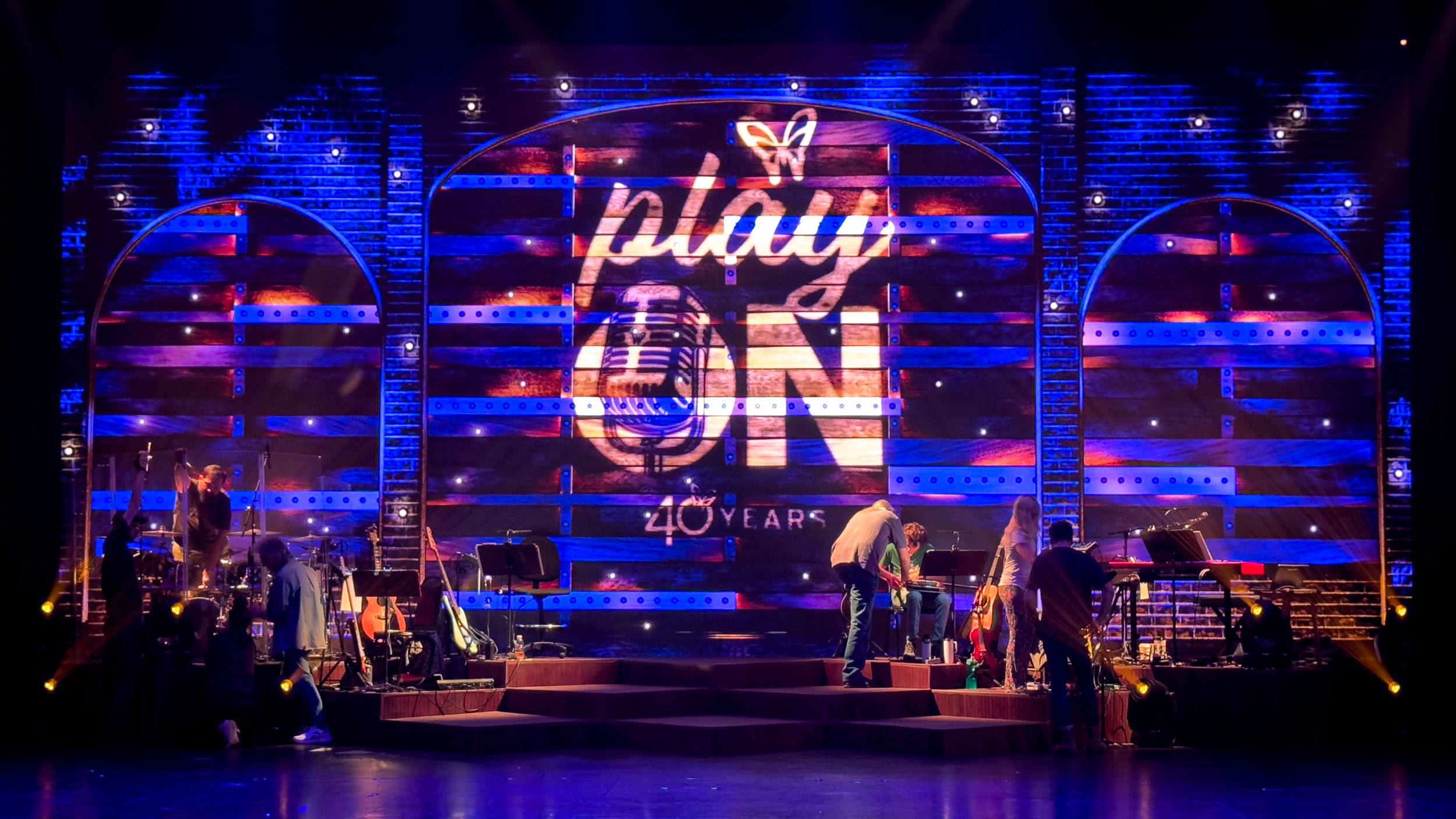 Stage with large screen displaying 'Play On, 40 Years', musical instruments and people preparing for a performance, colorful lighting with blue and purple tones.
