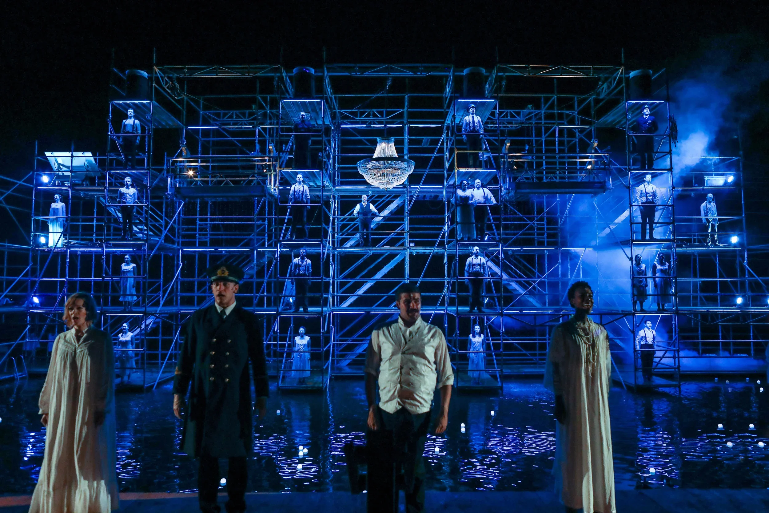 The stage of a theatrical production with actors in period costumes, featuring a large scaffolding structure with multiple levels and actors standing on different platforms. The scene is illuminated with blue lighting, with smoke effects and a chande