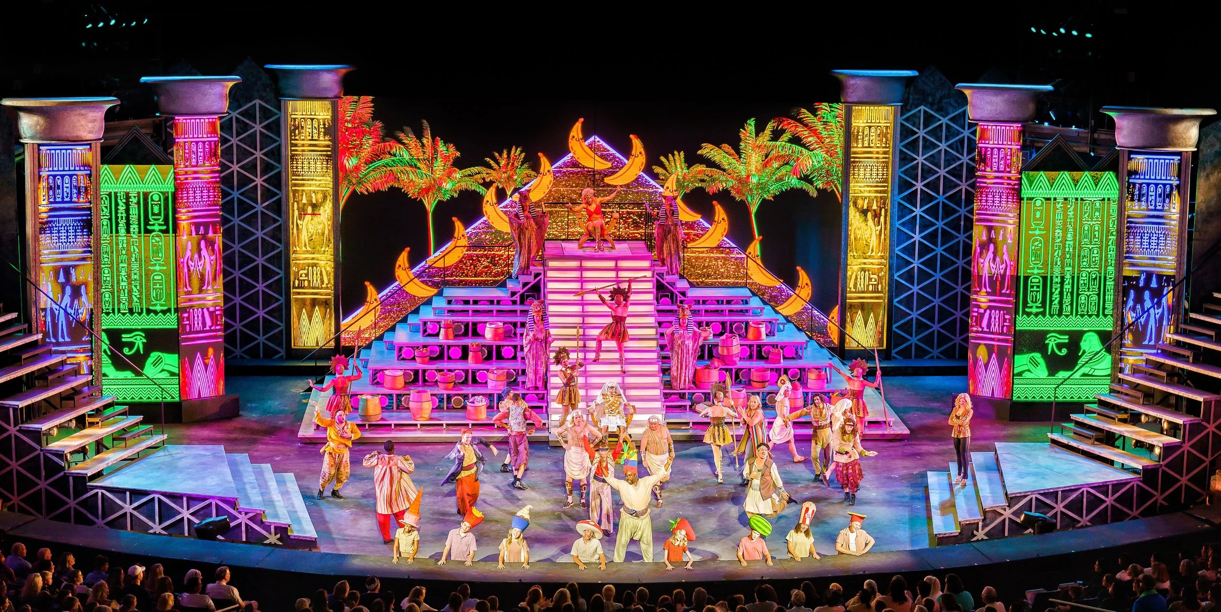 A colorful stage performance with vibrant costumes, set design featuring neon lights, tropical trees, and moon decorations, with performers dancing and singing in front of an audience.