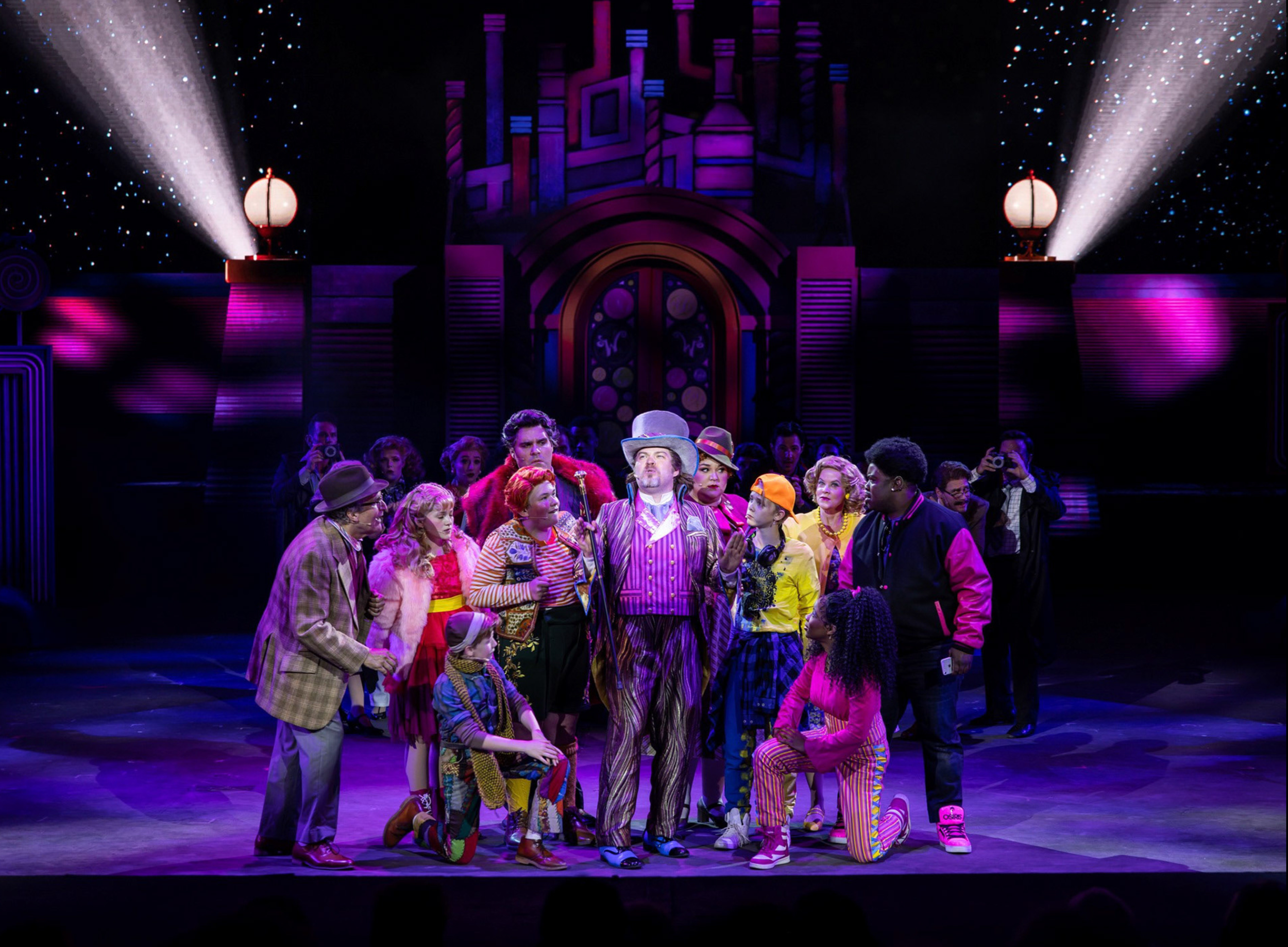 A theatrical scene with performers dressed in colorful costumes on stage, surrounded by an audience. The background features a stylized house with bright lighting and starry sky effects.