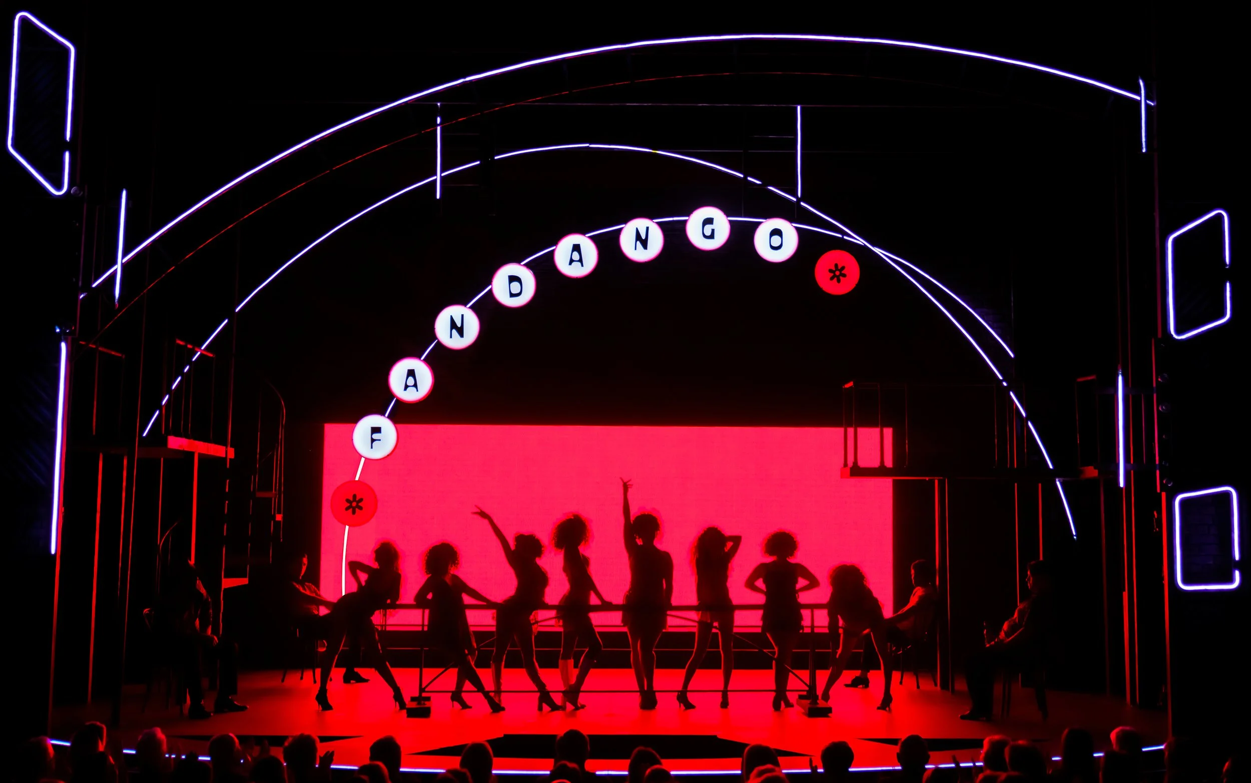Silhouettes of dancers on stage with a pink background, Neon signage spelling 'FANDANGO', and neon outlines of an arch and rectangles, with an audience watching in the foreground.