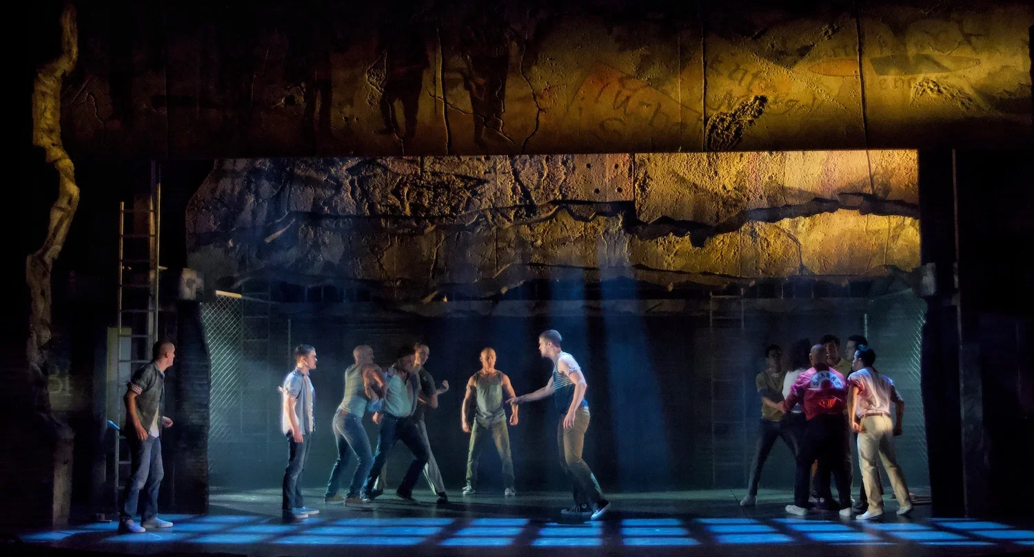 Theater scene with multiple actors on stage, some dancing and others standing, with a rocky backdrop and a blue-lit floor.