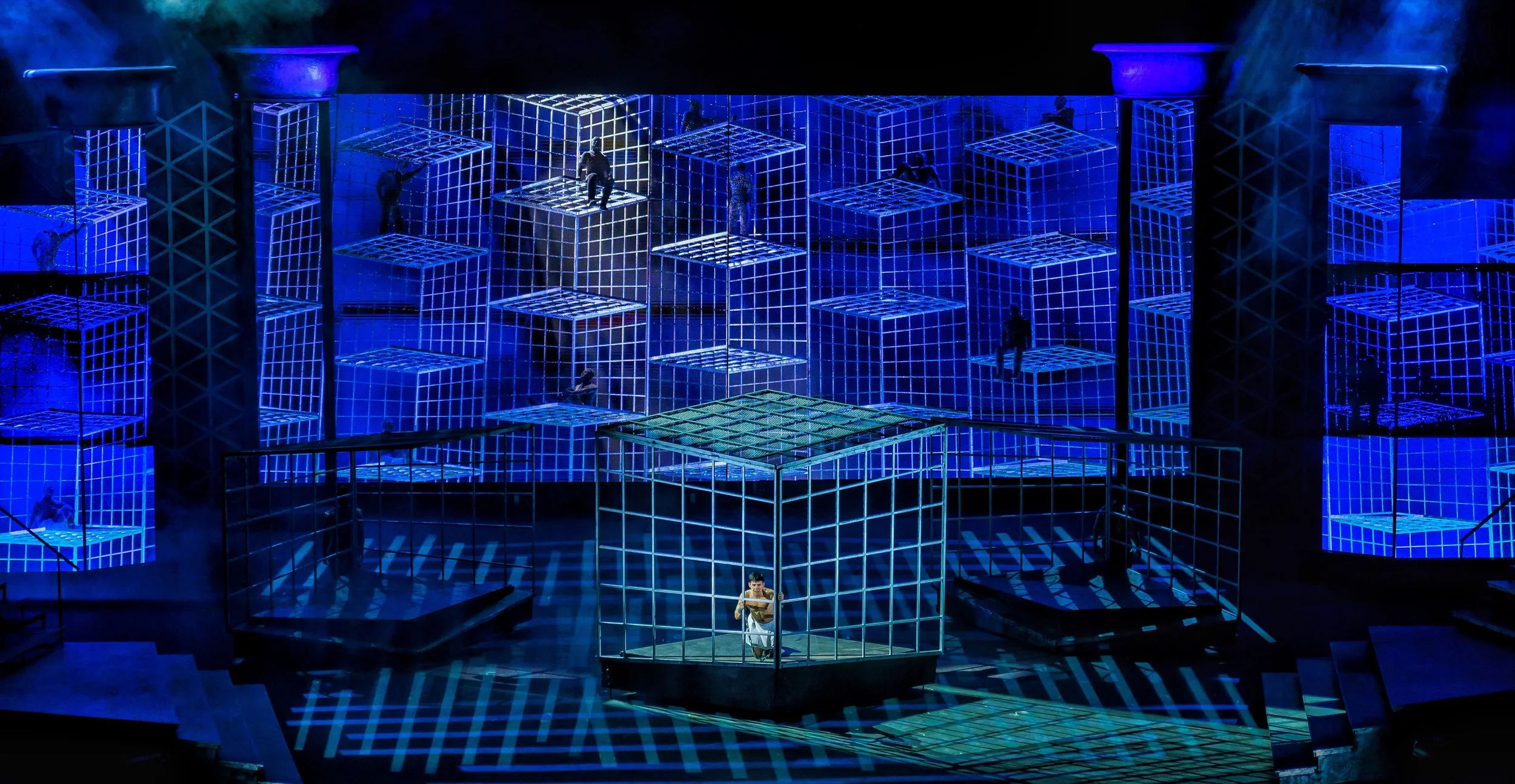 Stage scene with a large grid of illuminated blue cubes, some with performers inside, in a theatrical or concert setting.