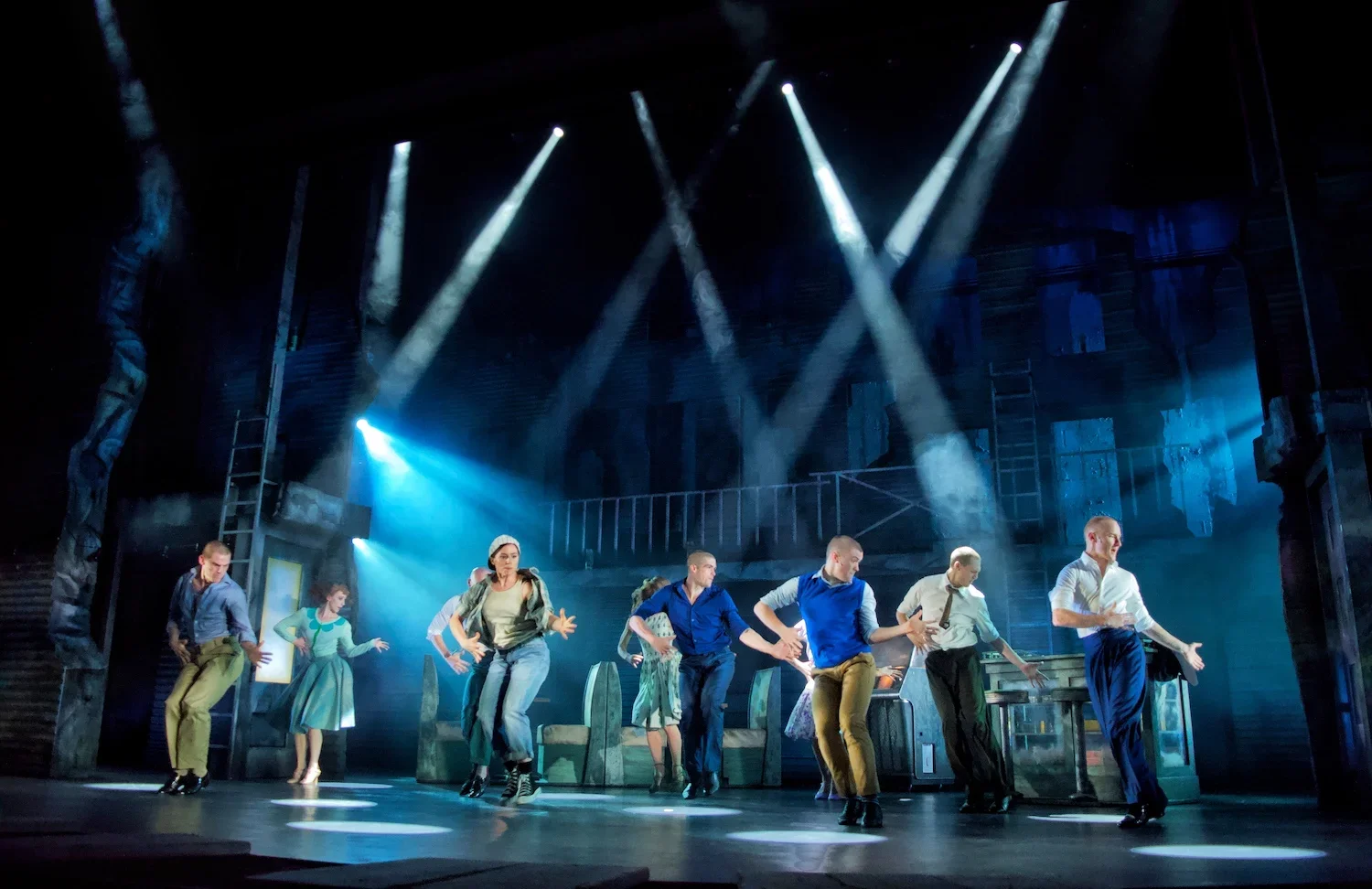 Theater stage with dancers performing under spotlights, with a set resembling an interior room with furniture and artwork.