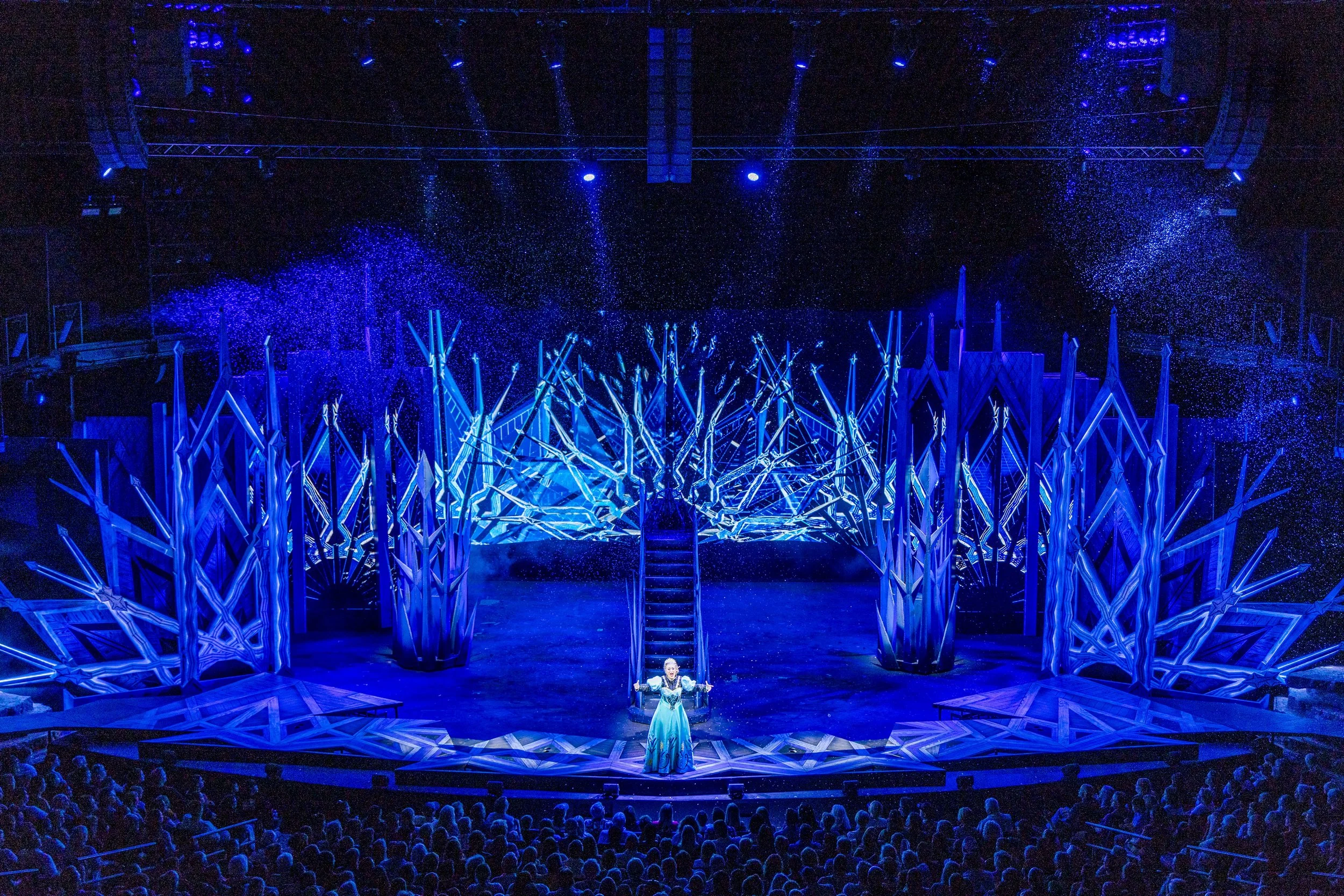 Stage set with blue lighting, snow-like particles, and a performer in a blue gown, audience in foreground.
