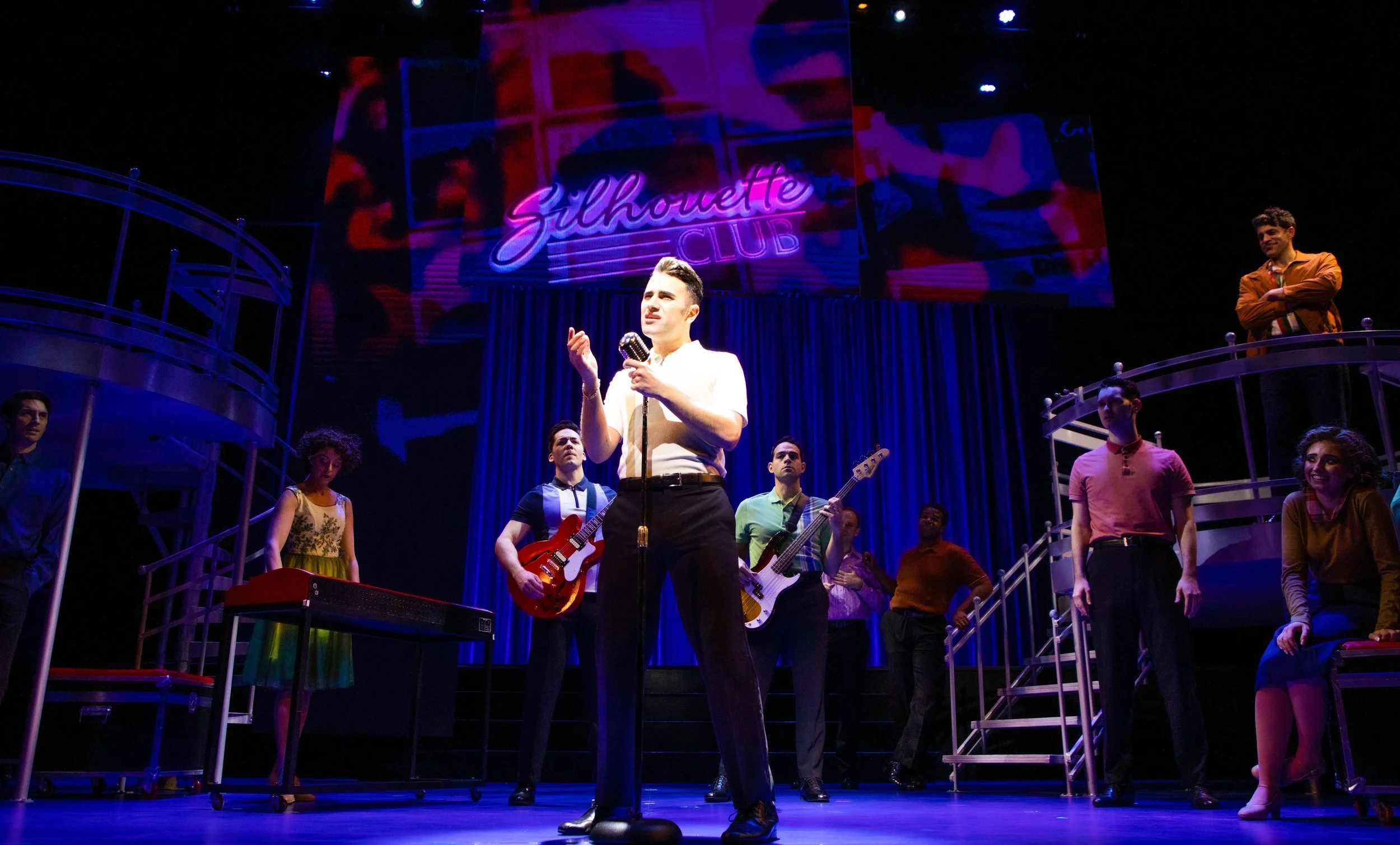 A stage scene from the musical 'Glamour Girls,' featuring a central male performer with a vintage microphone, surrounded by a diverse cast of actors. The backdrop displays a neon sign reading 'Glamour Club,' with vibrant lighting and a set that inclu