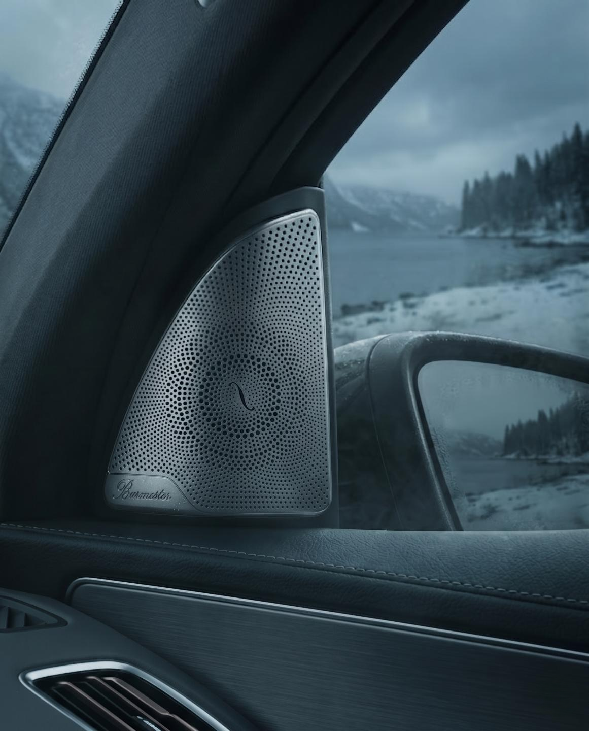 View from inside a car showing a Burmester speaker mounted on the A-pillar, with a snow-covered landscape, mountains, and trees visible outside through the windshield and side mirror.  Mercedes Eqc 400