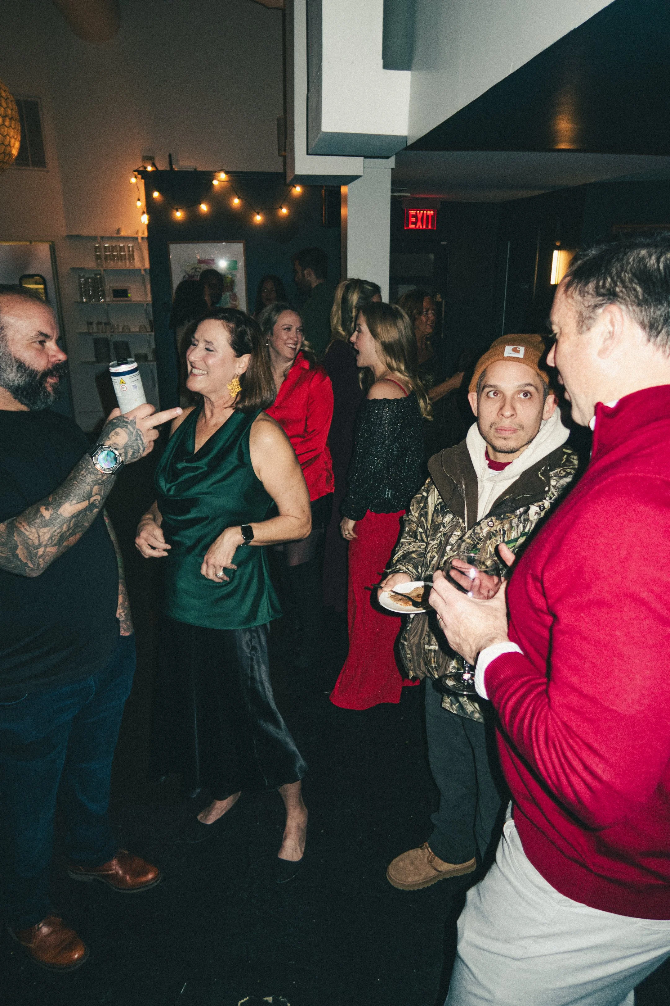 A group of people socializing at a party or gathering in a dimly lit indoor venue. Some are smiling and talking, holding drinks, with a background decorated with string lights and a few people visible in the background.