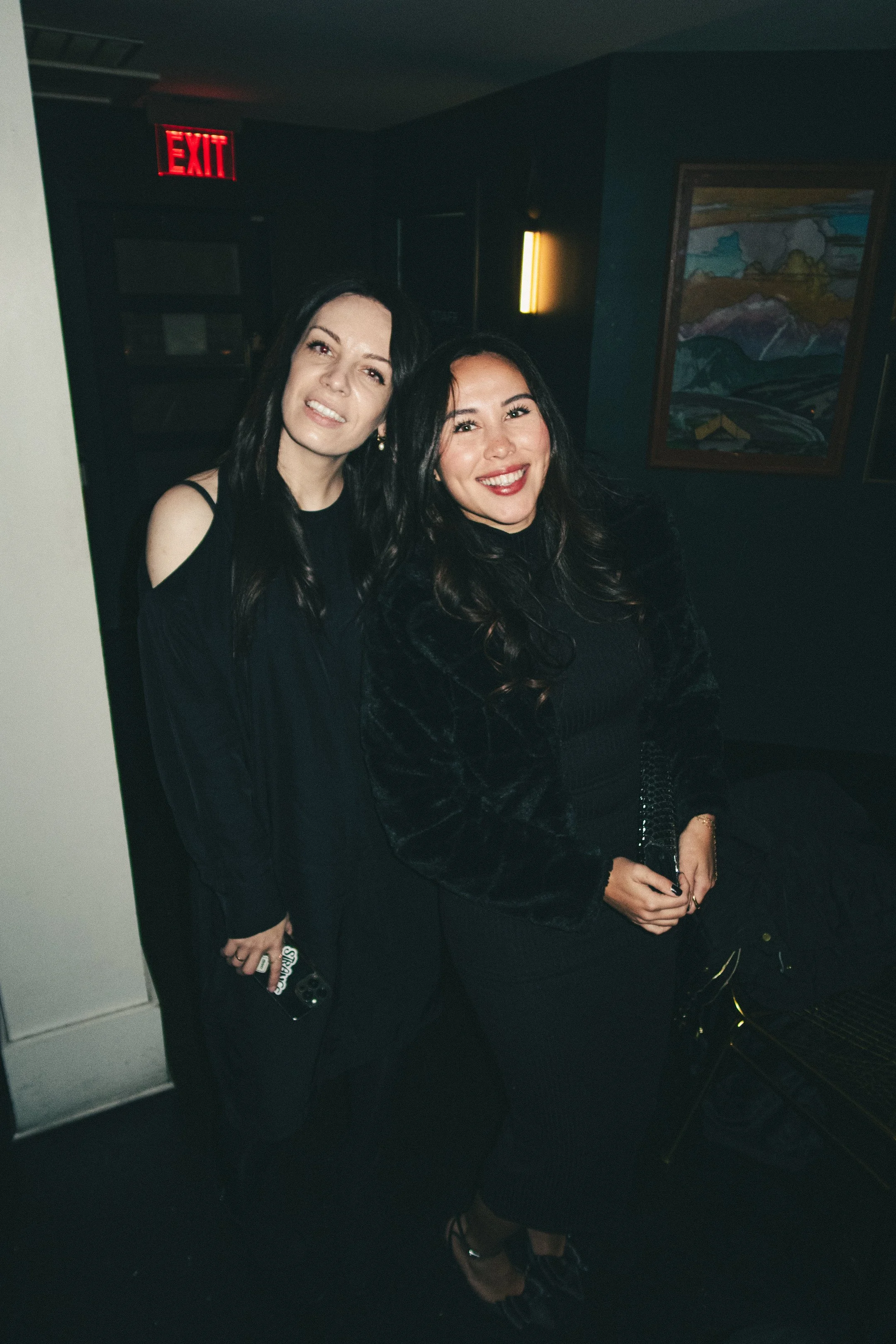 Two women with dark hair smiling and standing close together in a dimly lit room with dark walls, a colorful painting on the wall, and an illuminated exit sign above a doorway.